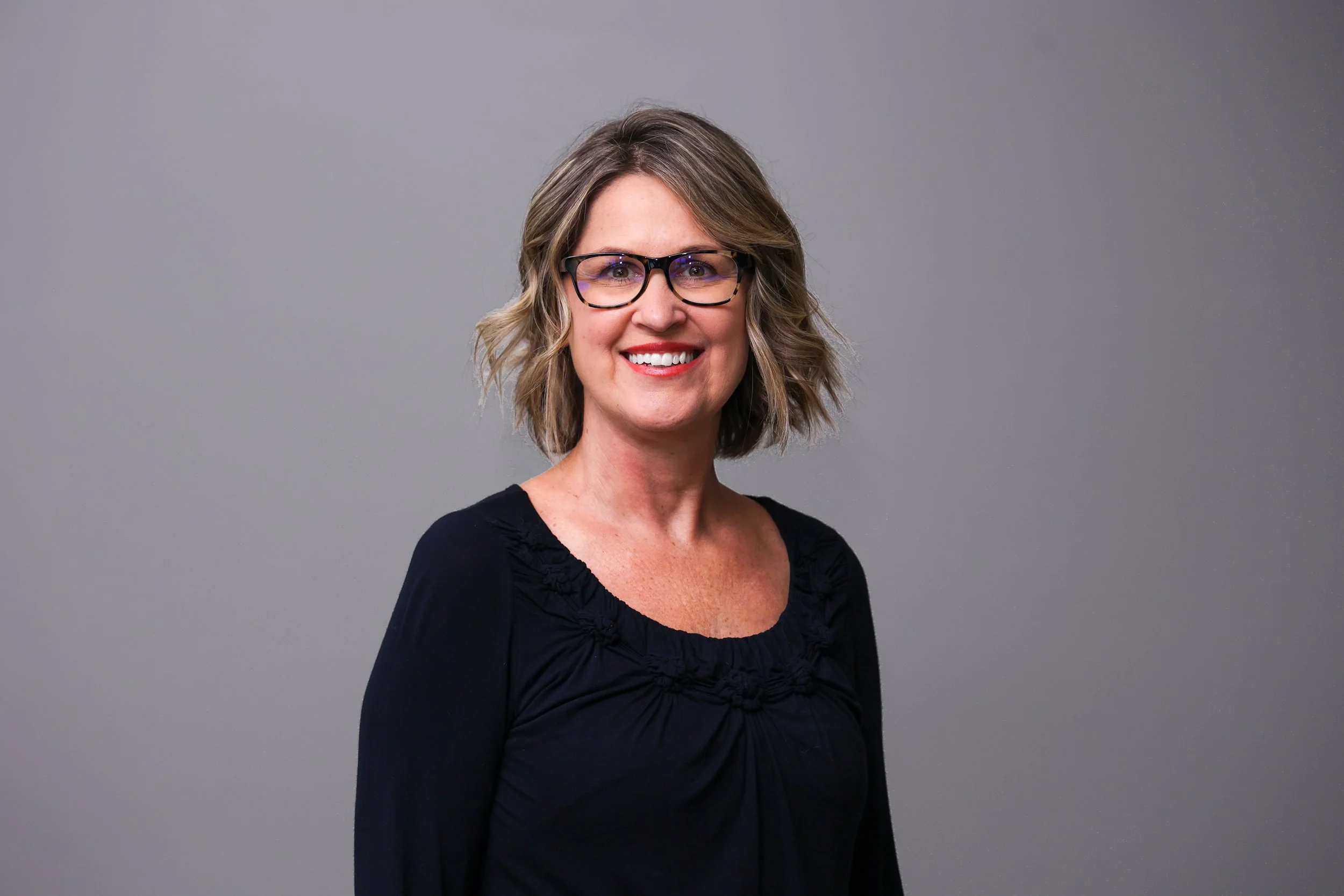A woman smiling, wearing glasses and a black-and-white striped shirt, standing in front of a wooden background.
