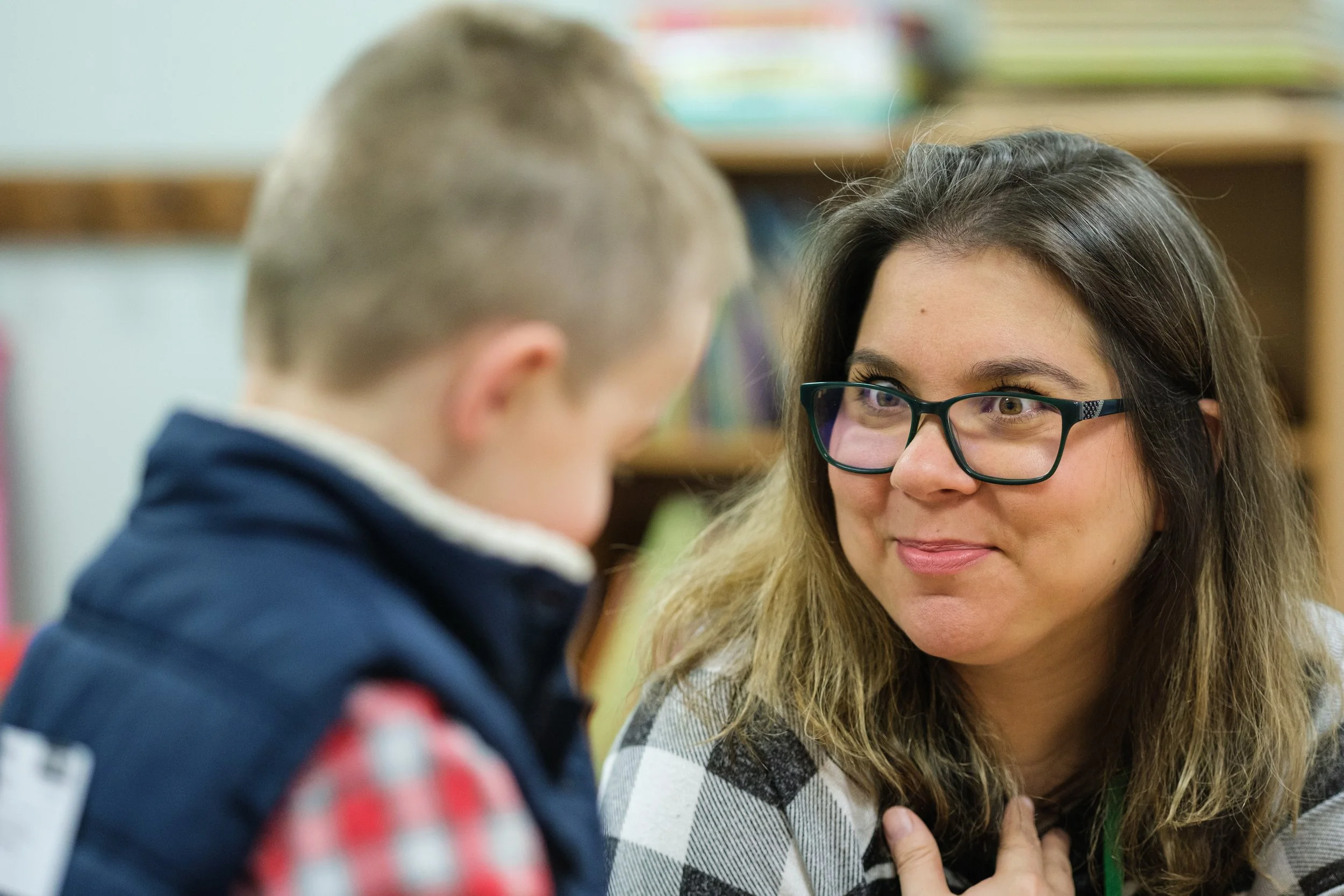 A woman with glasses and long hair smiling and looking at a young boy. The boy is facing away and has short light hair, wearing a dark jacket. They seem to be in a classroom or study environment with books on shelves in the background.