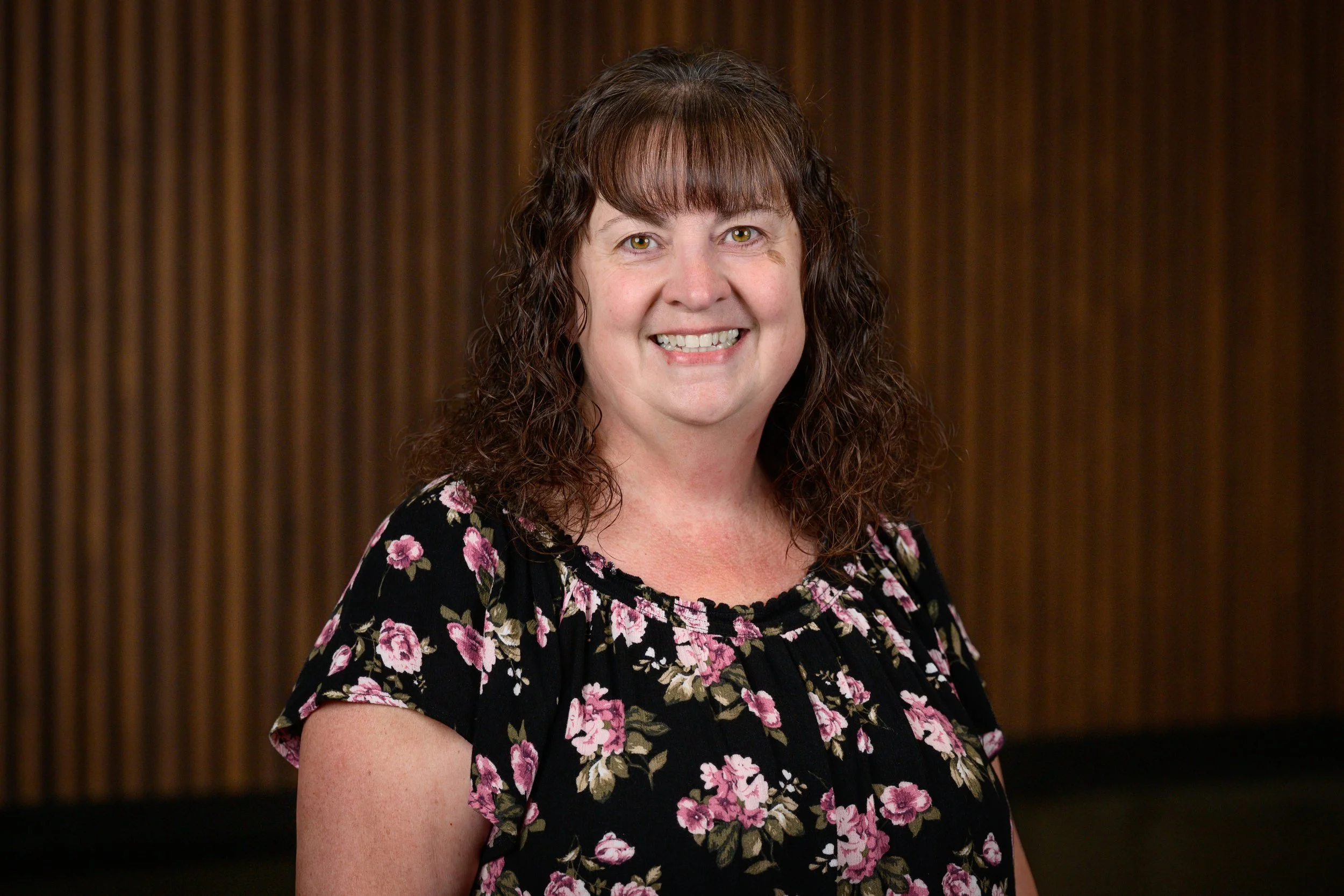 A smiling woman with curly brown hair and light skin wearing a black floral blouse stands in front of a brown wooden-paneled wall.