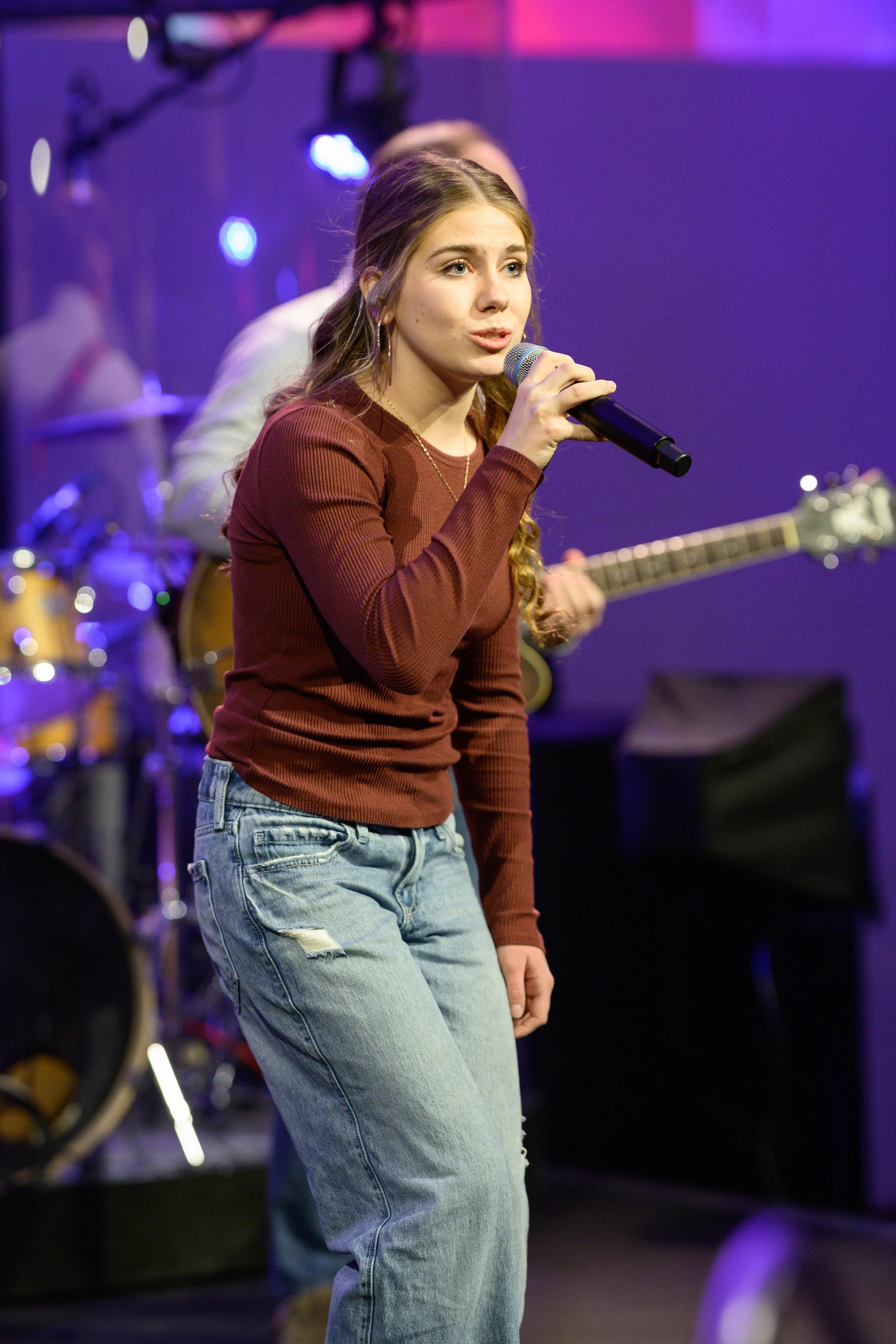 Young woman singing into a microphone during a live music performance, with a guitarist and drums in the background, on a stage with purple and pink lighting.