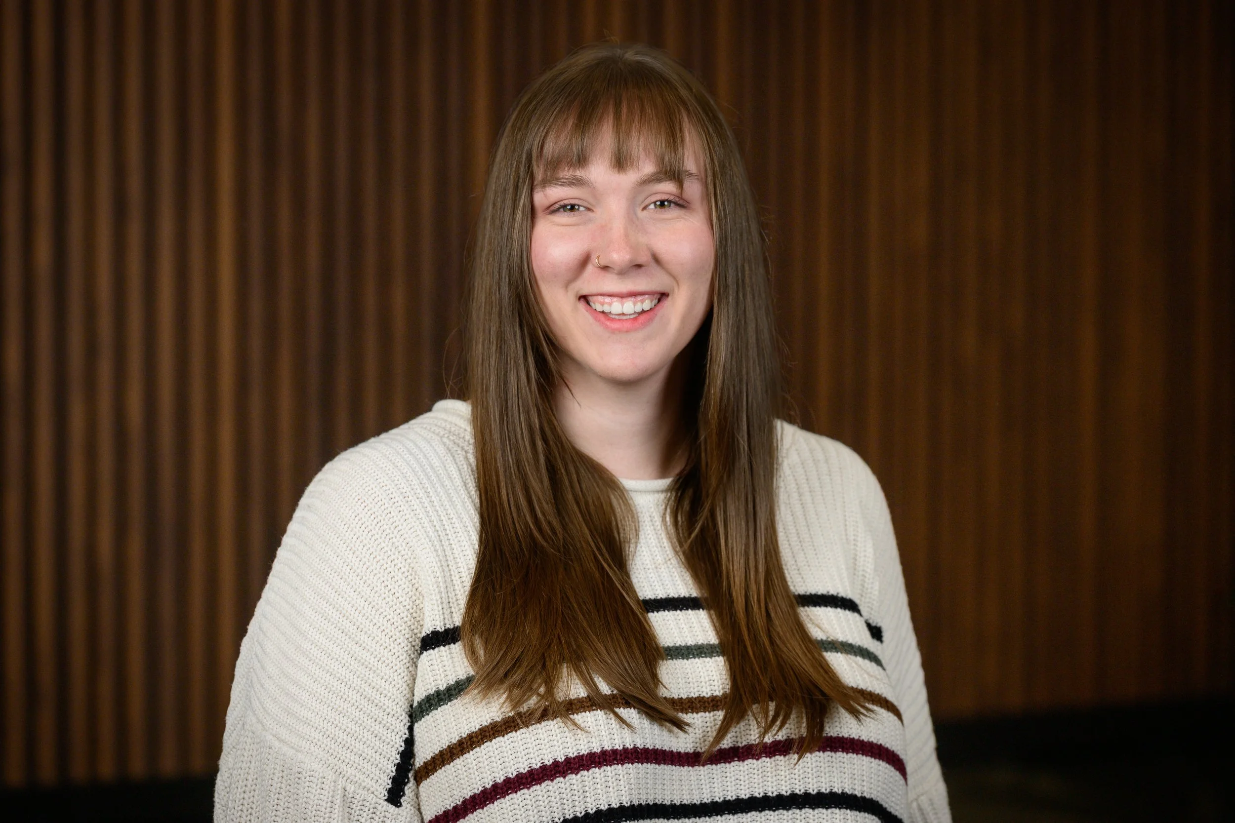 Young woman with long brown hair, wearing a cream and striped sweater, smiling in front of a wooden background.