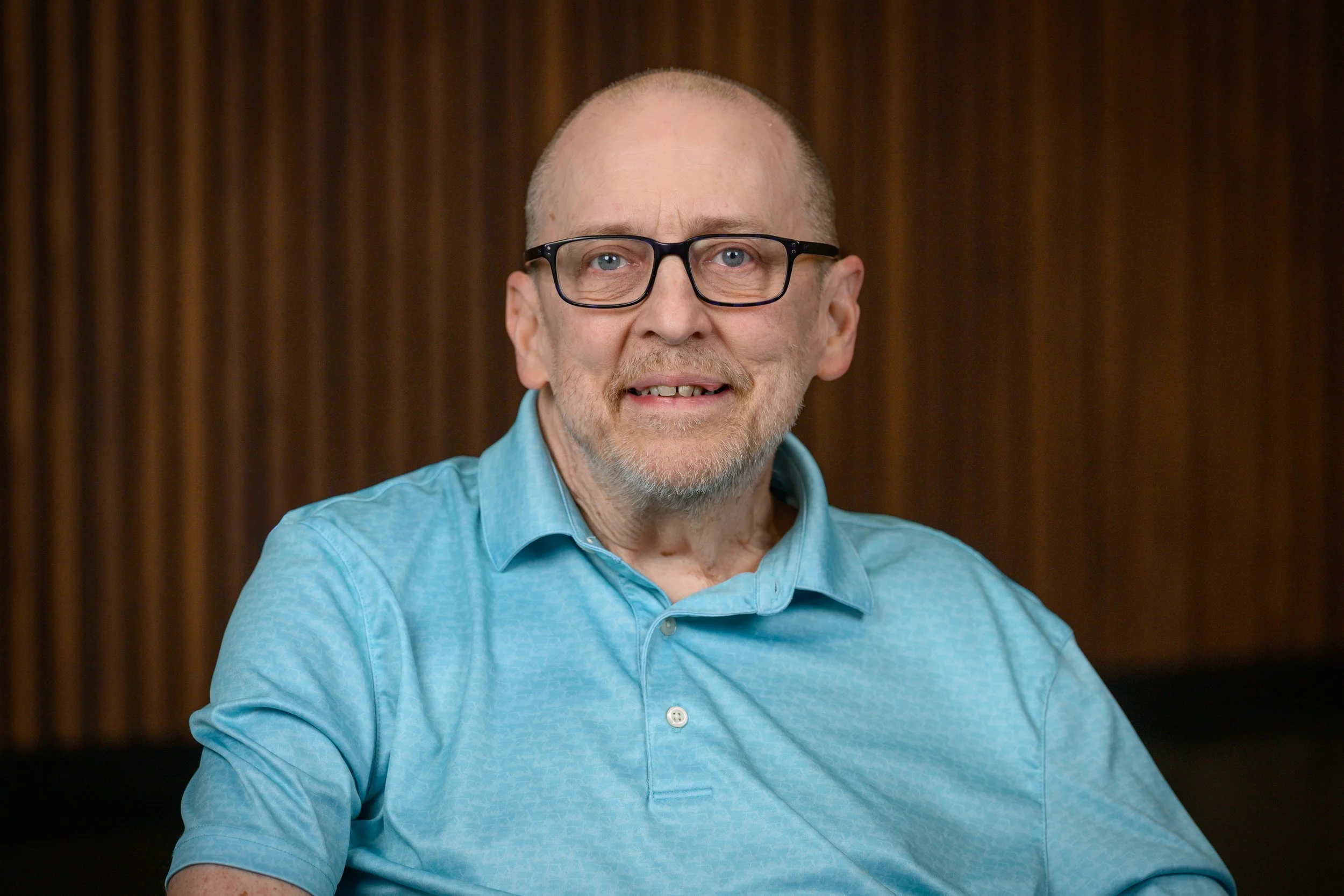 A middle-aged man with glasses, a short beard, wearing a blue button-up shirt, smiling, and sitting in front of a dark wooden background.