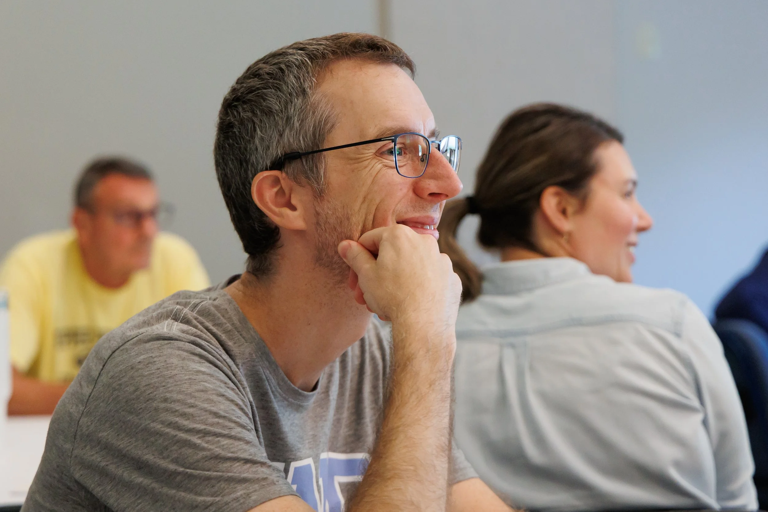 A man with glasses smiling and resting his chin on his hand, attending a meeting with other people, including a woman and a man, in a conference room.