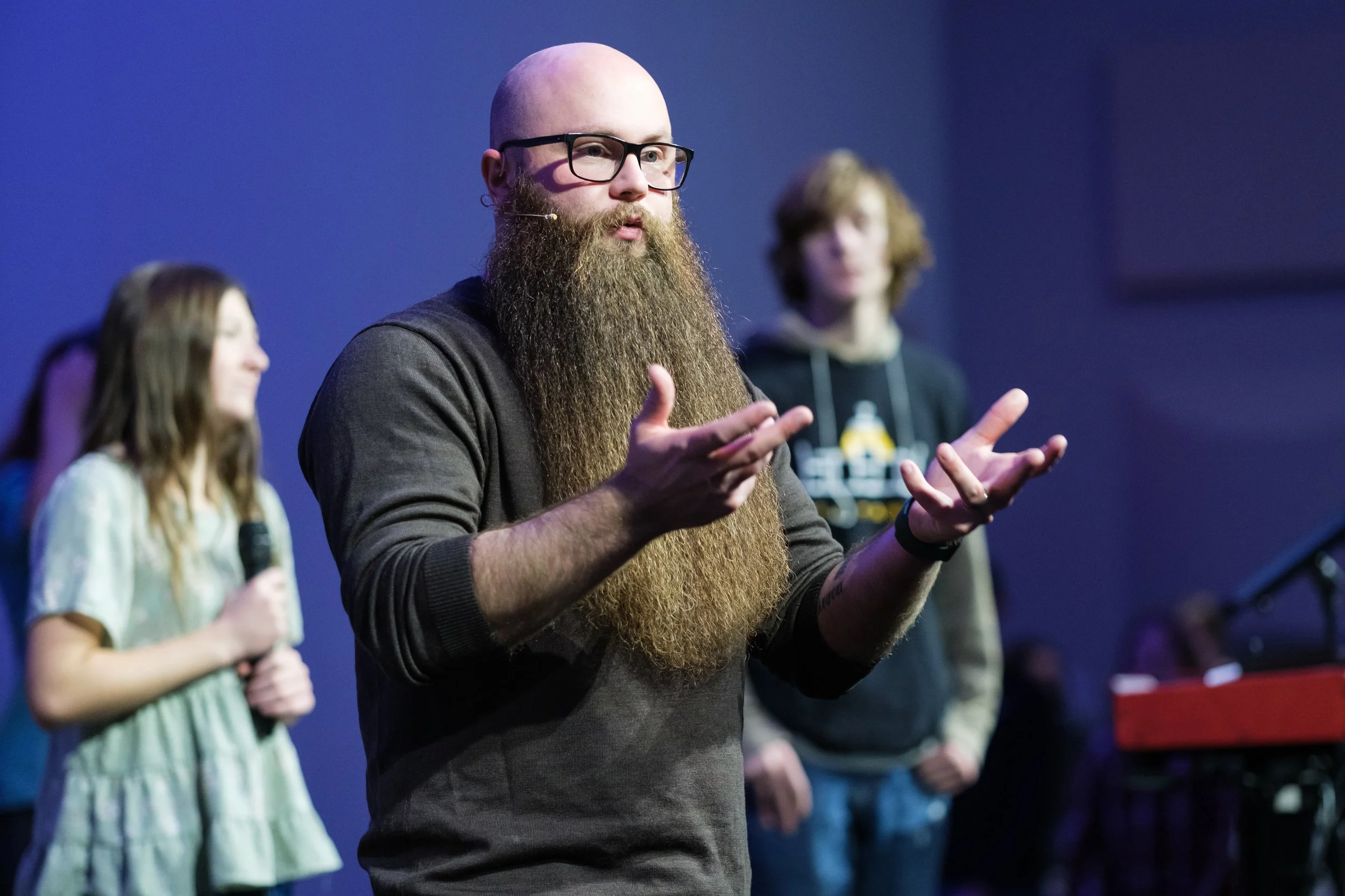A group of people on stage, with one man in the foreground speaking and gesturing with his hands, wearing glasses and a beard, while a woman with long hair and a microphone stands to the left, and others are visible in the background.