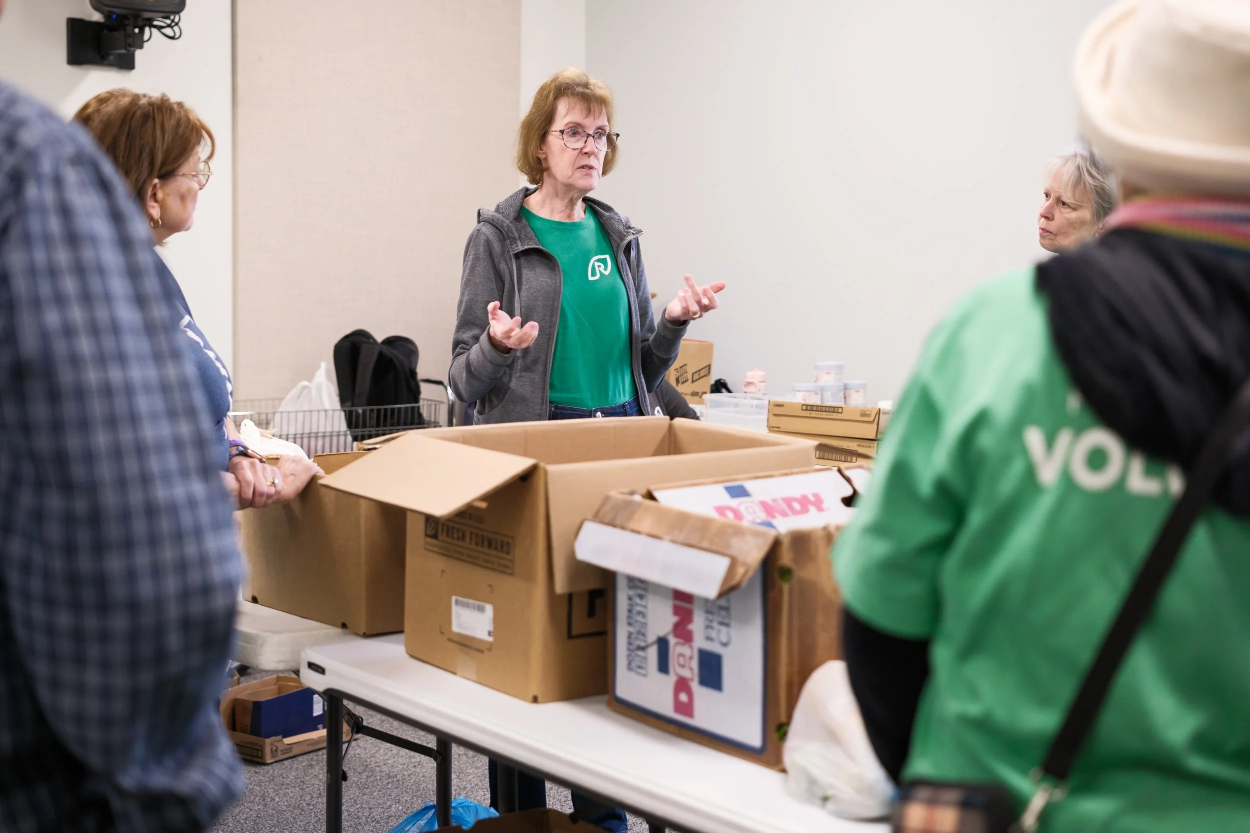 A woman in a green shirt and glasses is standing behind a table with cardboard boxes and talking to a small group of people in a room. The room appears to be a volunteer or community center, with boxes and supplies on the table.