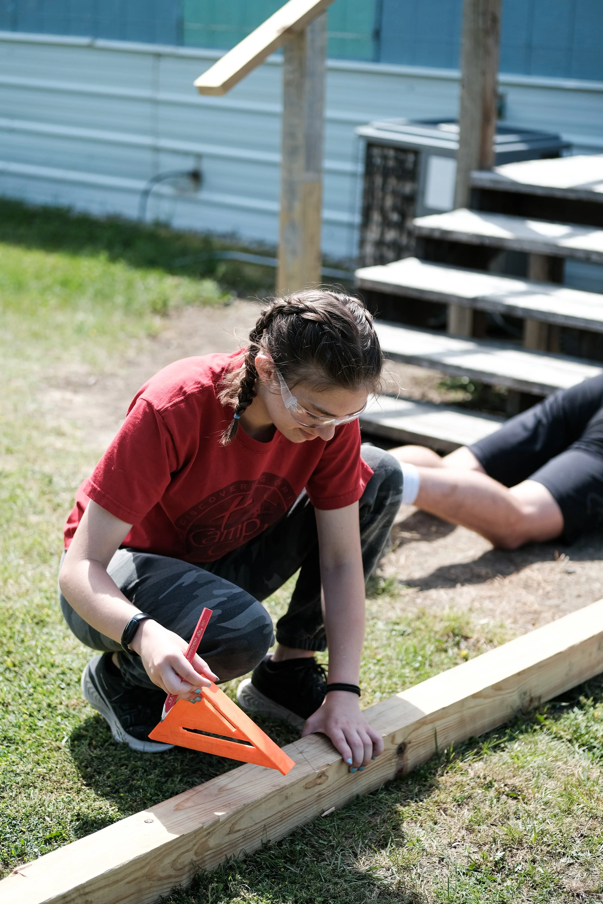 A young girl with glasses and braided hair kneeling on grass while measuring and marking a wooden plank with a pencil and carpenter's square outdoors, with a blue house, stairs, and a gray air conditioning unit in the background.