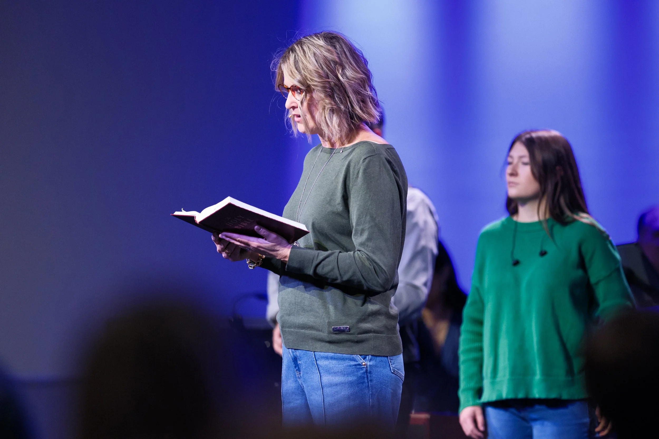A woman with curly blonde hair, wearing glasses, a green long-sleeve top, and blue jeans, is reading from a black book on stage, with other people standing in the background under purple and blue stage lighting.