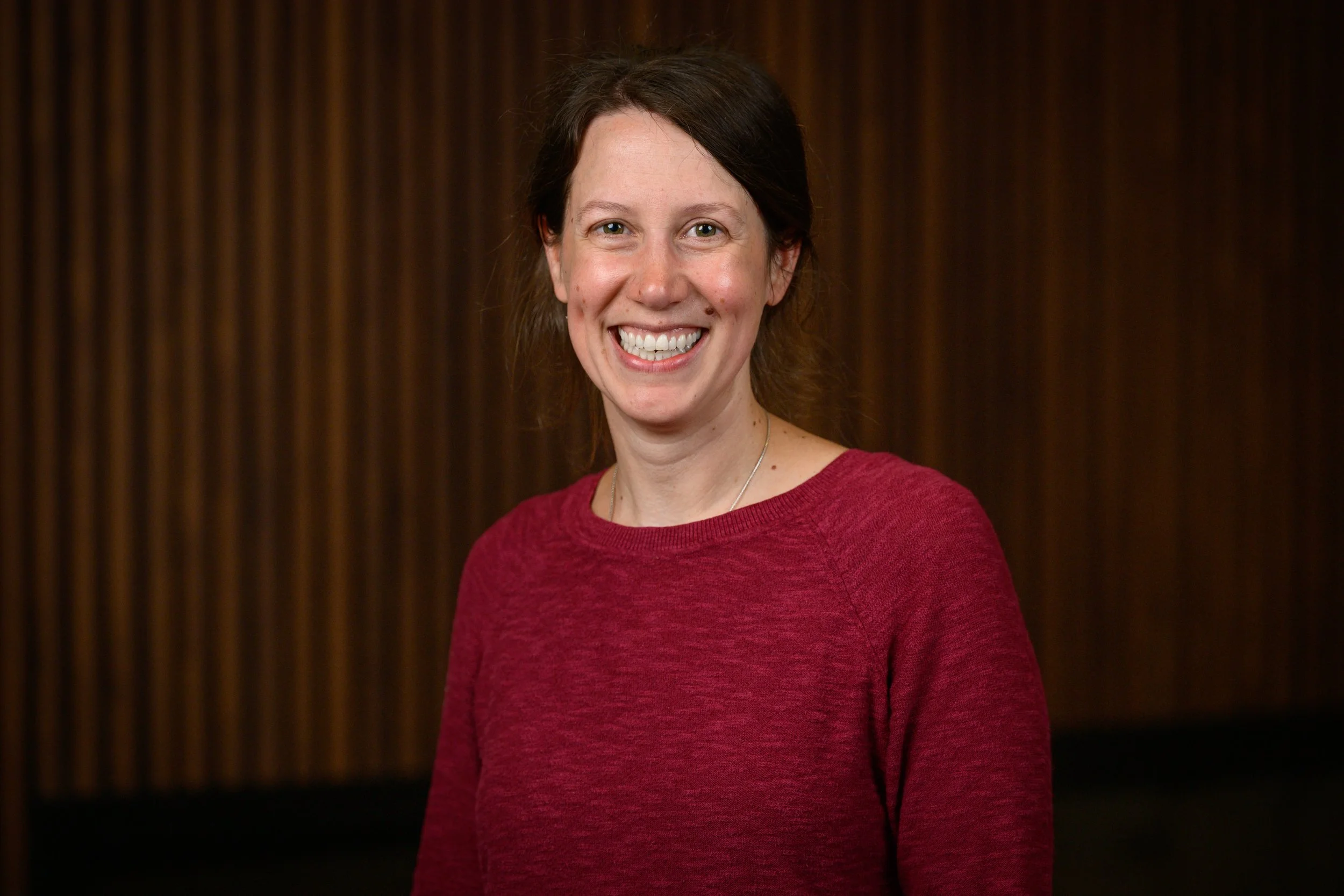 A smiling woman with dark hair wearing a red sweater standing in front of a dark wooden background.