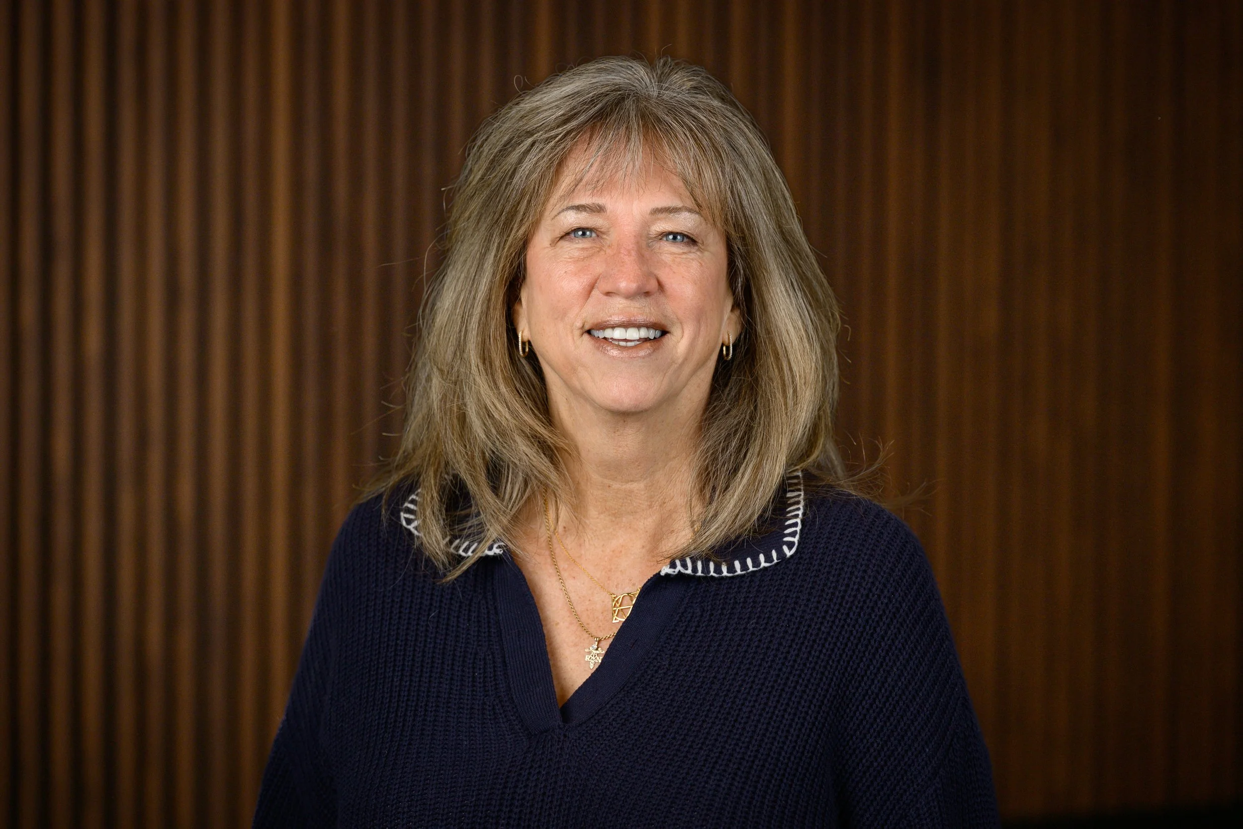 A smiling woman with shoulder-length blonde hair, wearing a navy blue sweater with white trim and a gold necklace, standing against a wooden-paneled background.