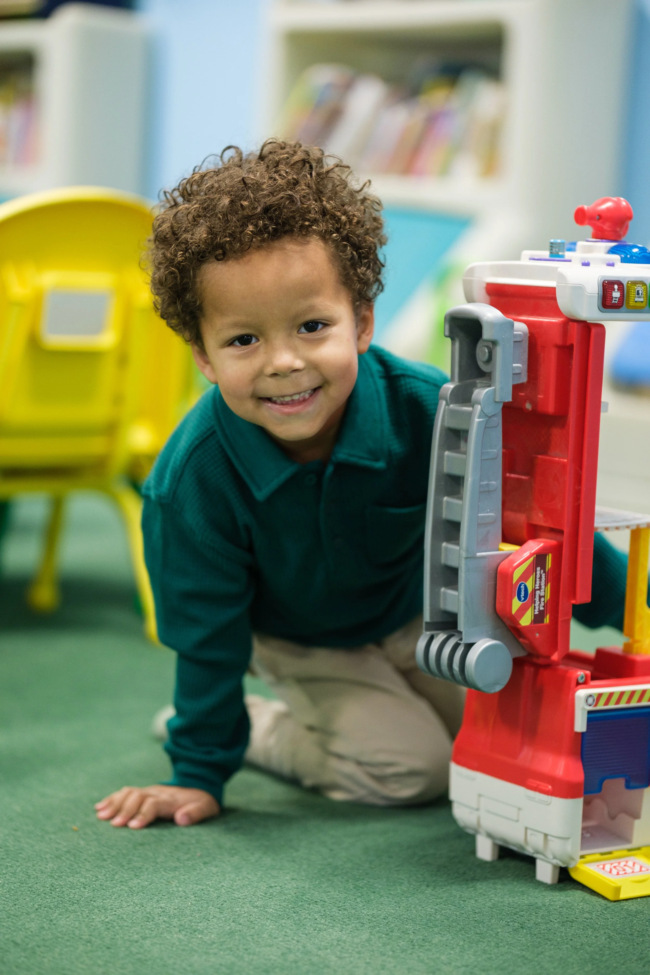 A young boy with curly hair smiling while crawling on a green carpet in a playroom. There are yellow chairs and bookshelves with books in the background. A toy parking garage is partially visible to the right.