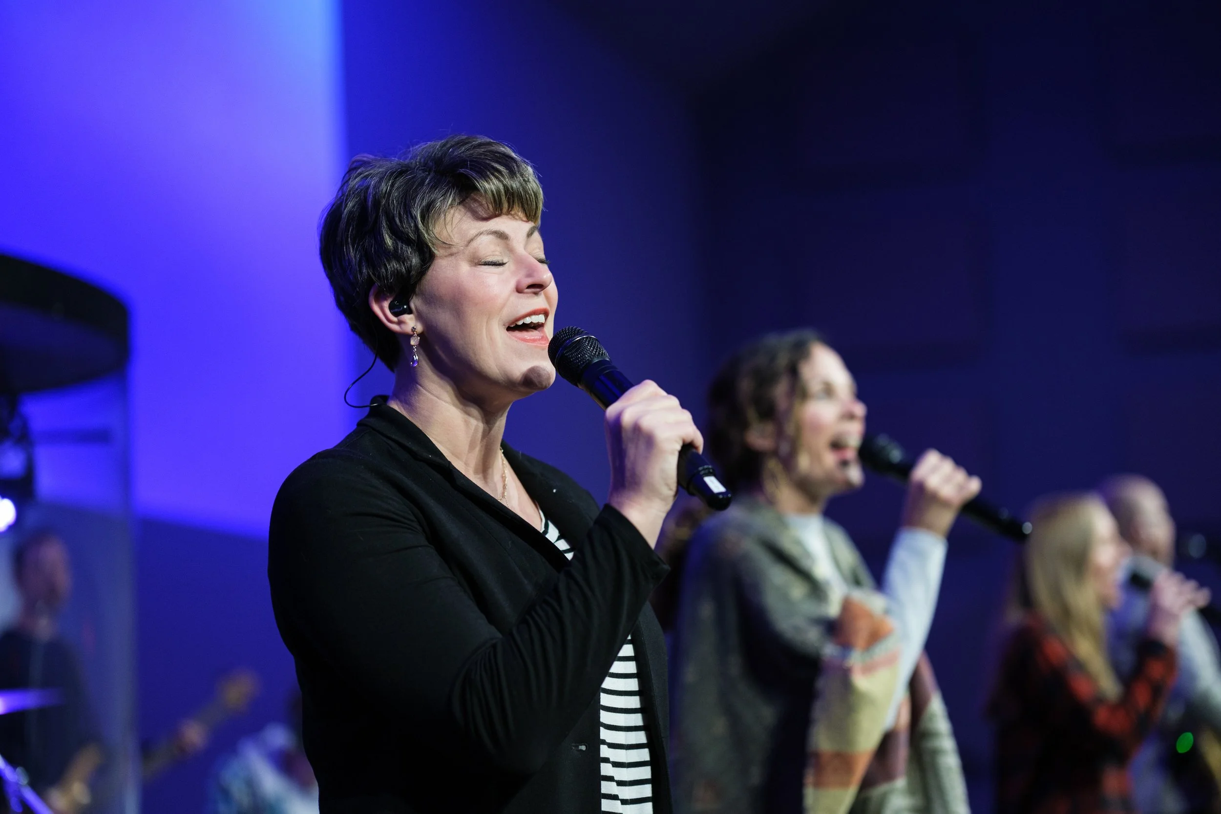 A woman with short hair singing into a microphone during a group performance, with other women in the background also singing.