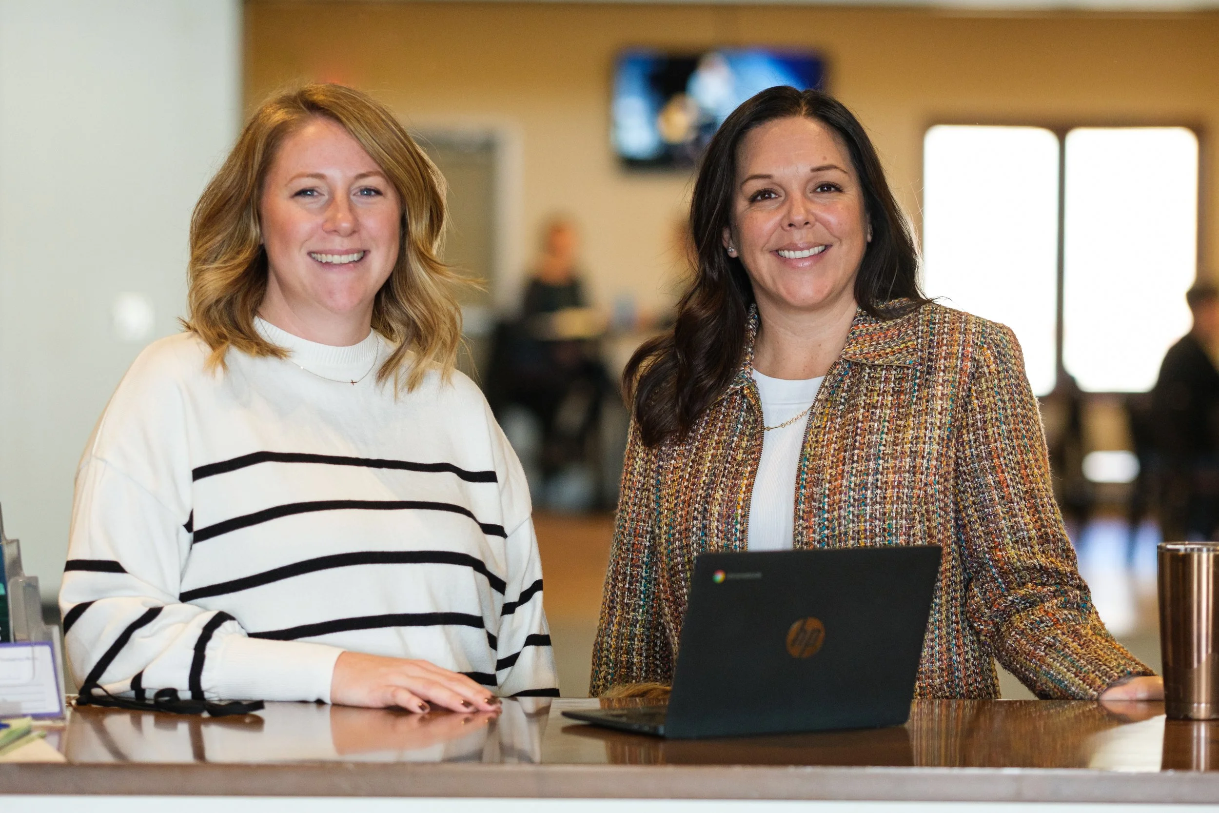 Two women standing at a counter in an indoor setting, smiling at the camera. One woman has shoulder-length blond hair and is wearing a white and black striped sweater. The other woman has long dark hair and is wearing a colorful jacket over a white top. There is a laptop in front of the woman with dark hair, and the background shows a blurred indoor space with windows and other people.