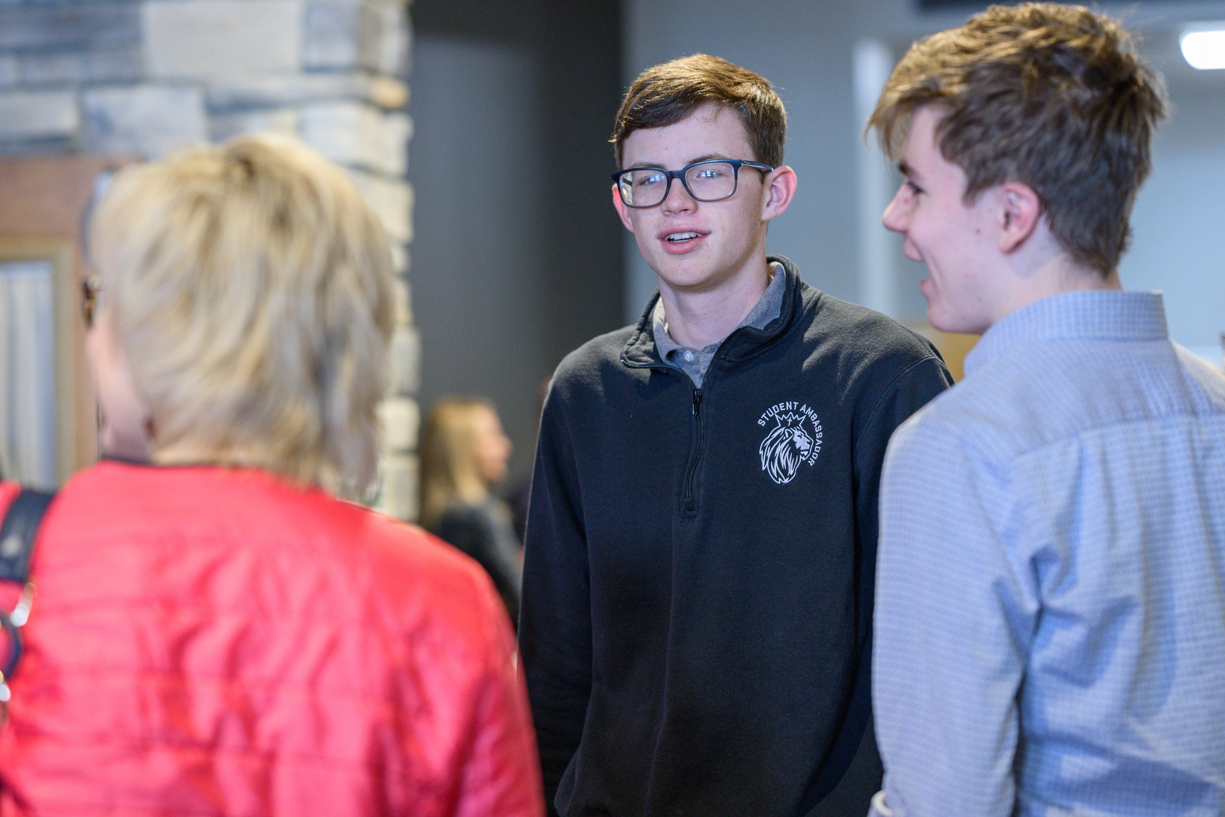 Three young people engaged in conversation indoors. One young man with glasses and a black jacket is speaking, while another young man and an older woman with blonde hair are listening.