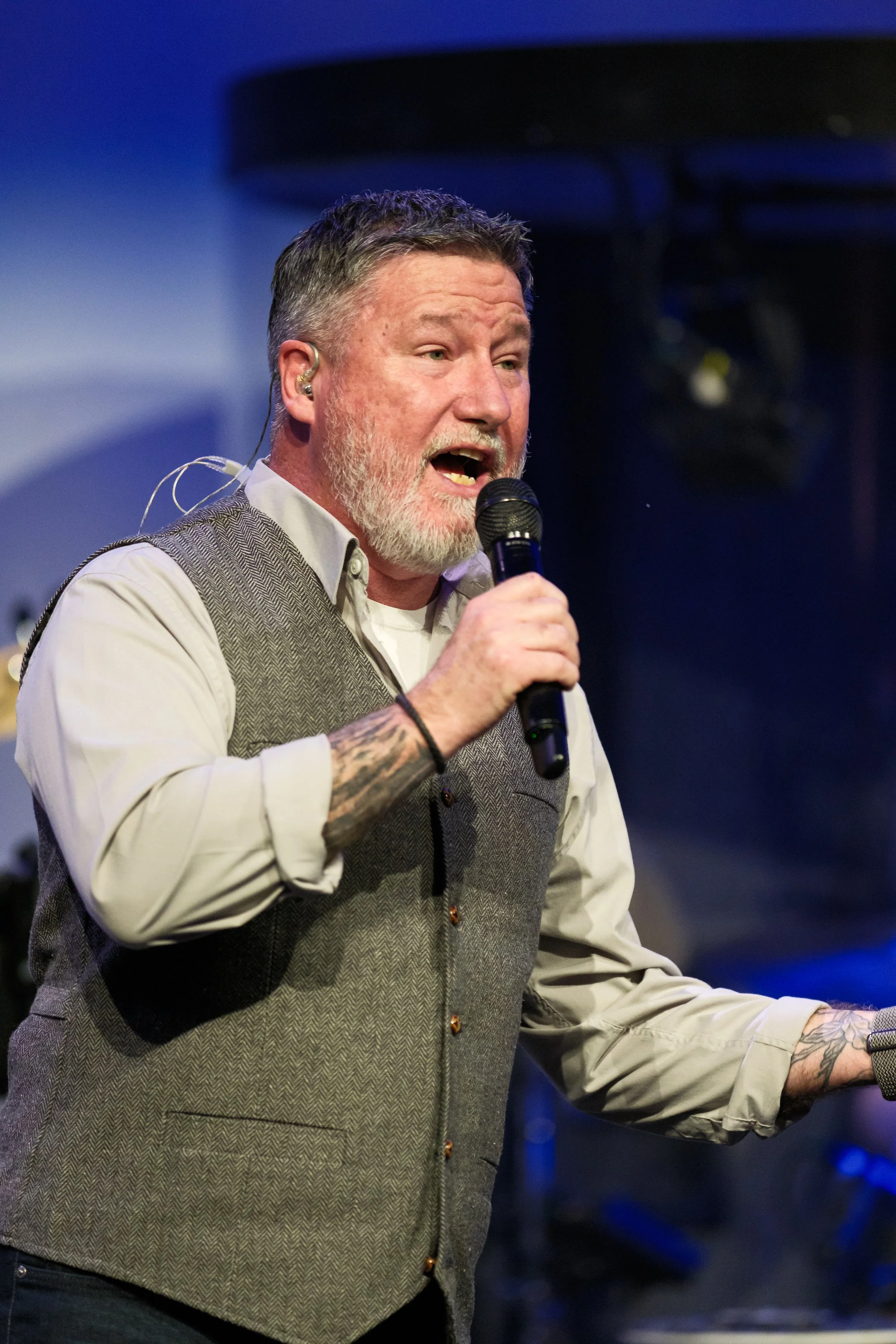 A man with gray hair and beard singing into a microphone during a performance, wearing a beige shirt and gray vest, tattooed on one arm, with a dark background and stage lighting.