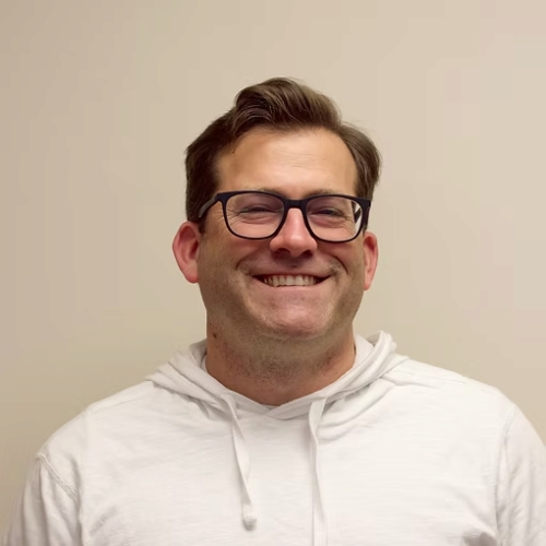 A smiling man with glasses wearing a white hoodie, standing against a plain beige background.