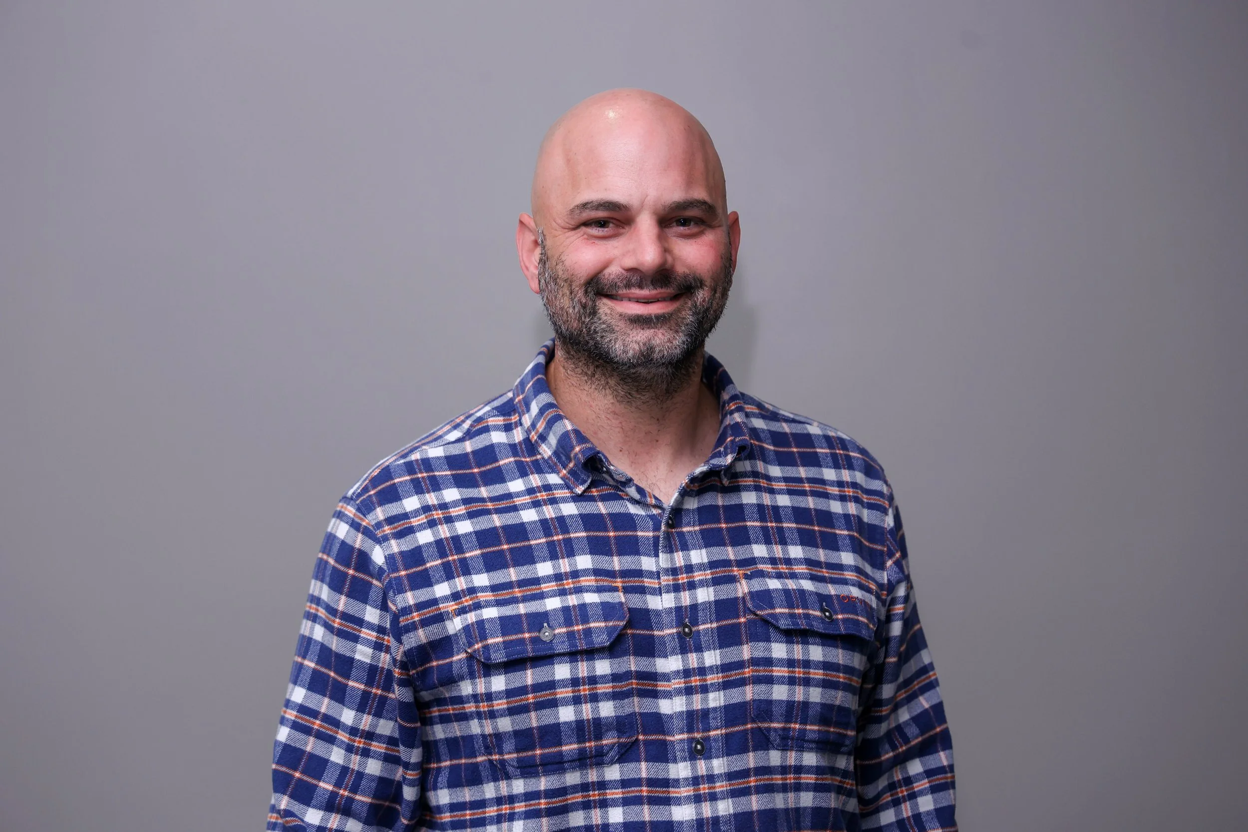 A smiling bald man with a beard, wearing a blue and orange plaid shirt, standing against a plain gray background.