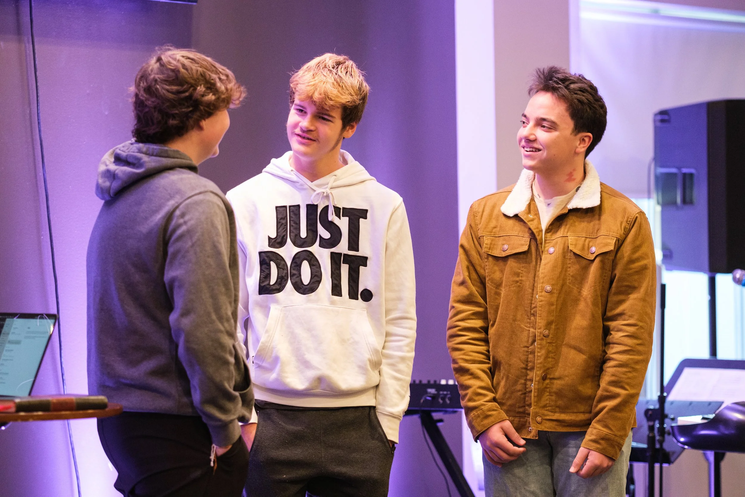 Three teenage boys talking in a room with purple lighting, one wearing a white hoodie with 'Just Do It' text, another in a brown jacket with a shearling collar, and the third facing away wearing a gray hoodie.