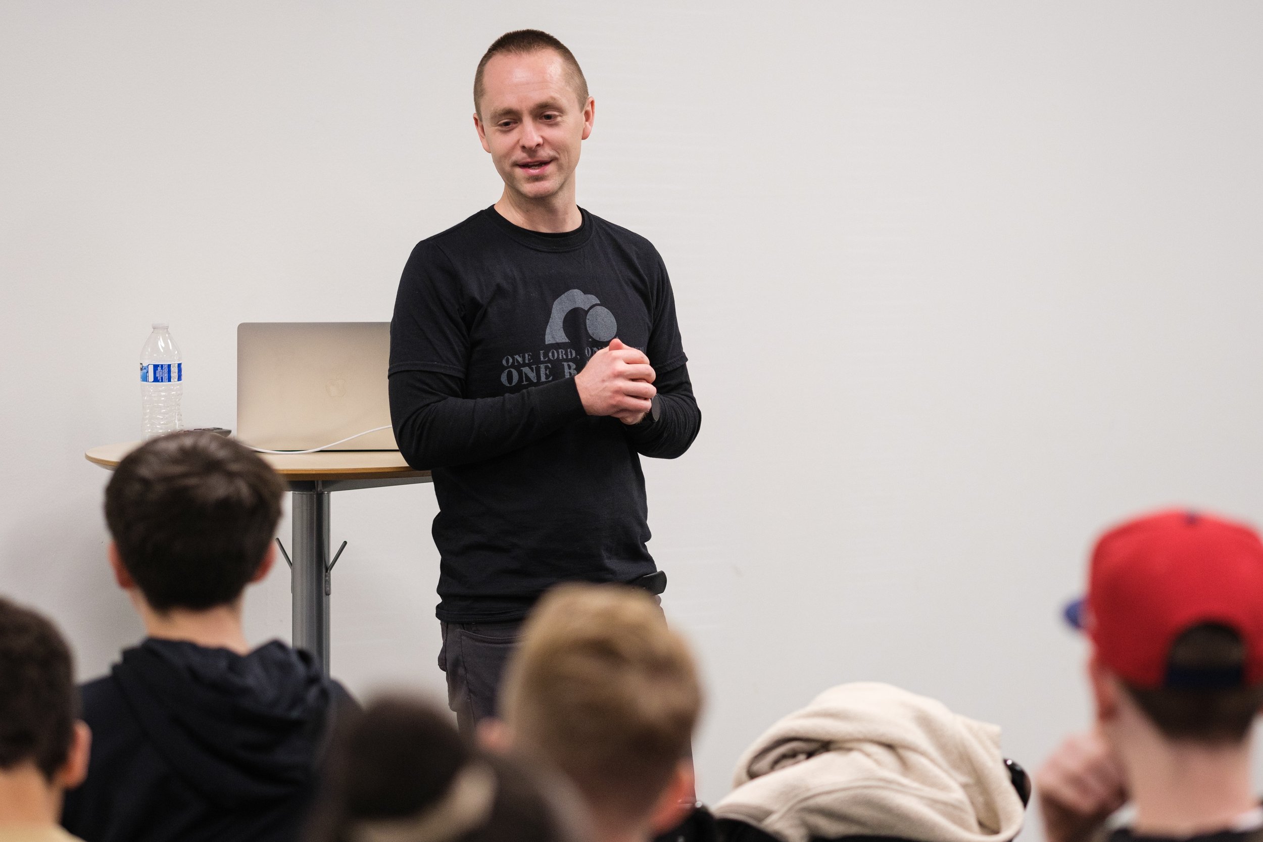 A man stands in front of a group, speaking or presenting, in a room with a white wall. He is wearing a black t-shirt with text and a logo, with his hands clasped together. Several people are sitting and listening, some with their backs to the camera.
