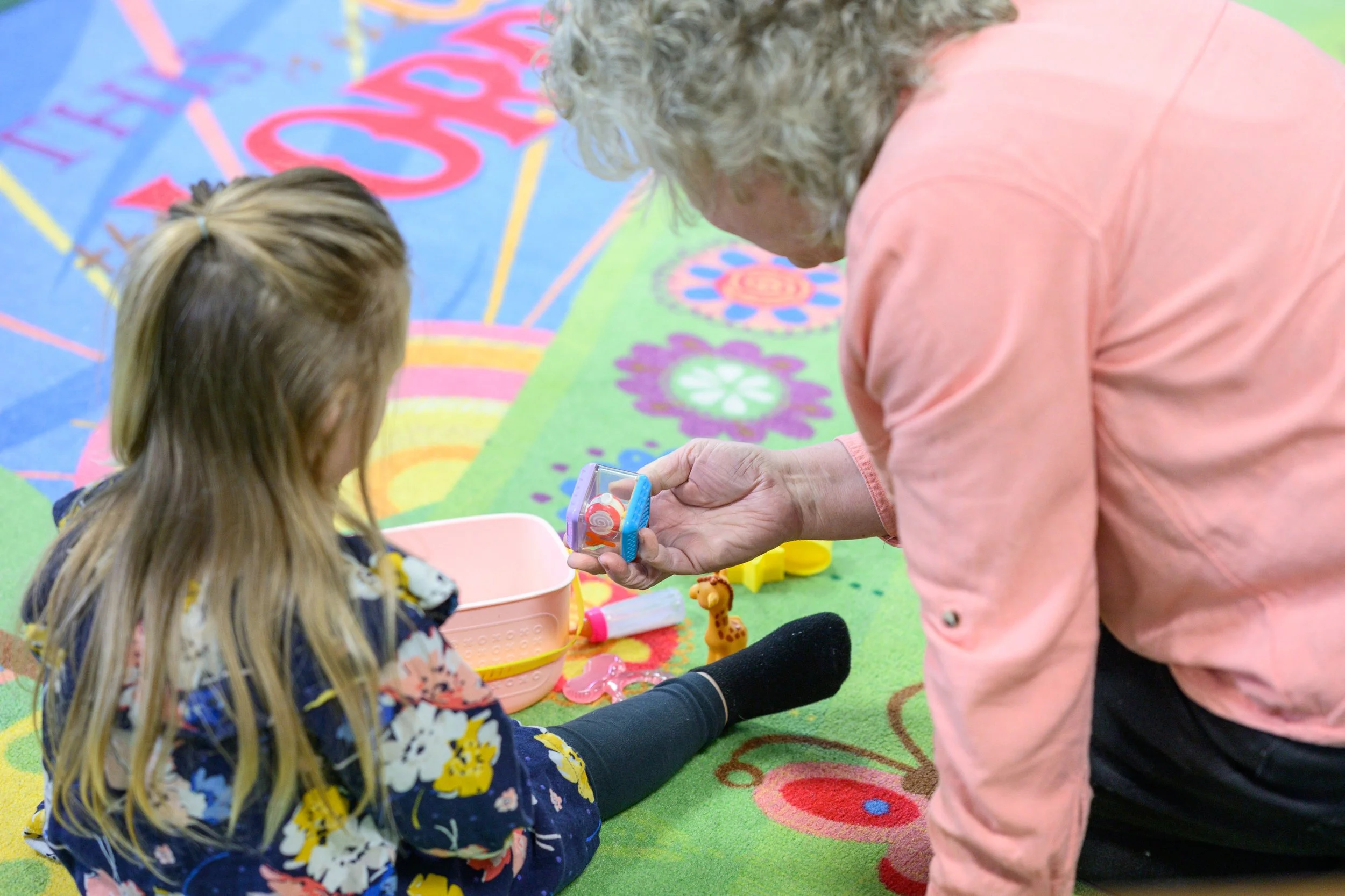 An elderly woman and a young girl are playing together on a colorful play mat indoors. The woman is holding a toy while the girl, with long blonde hair tied back, sits on the mat.