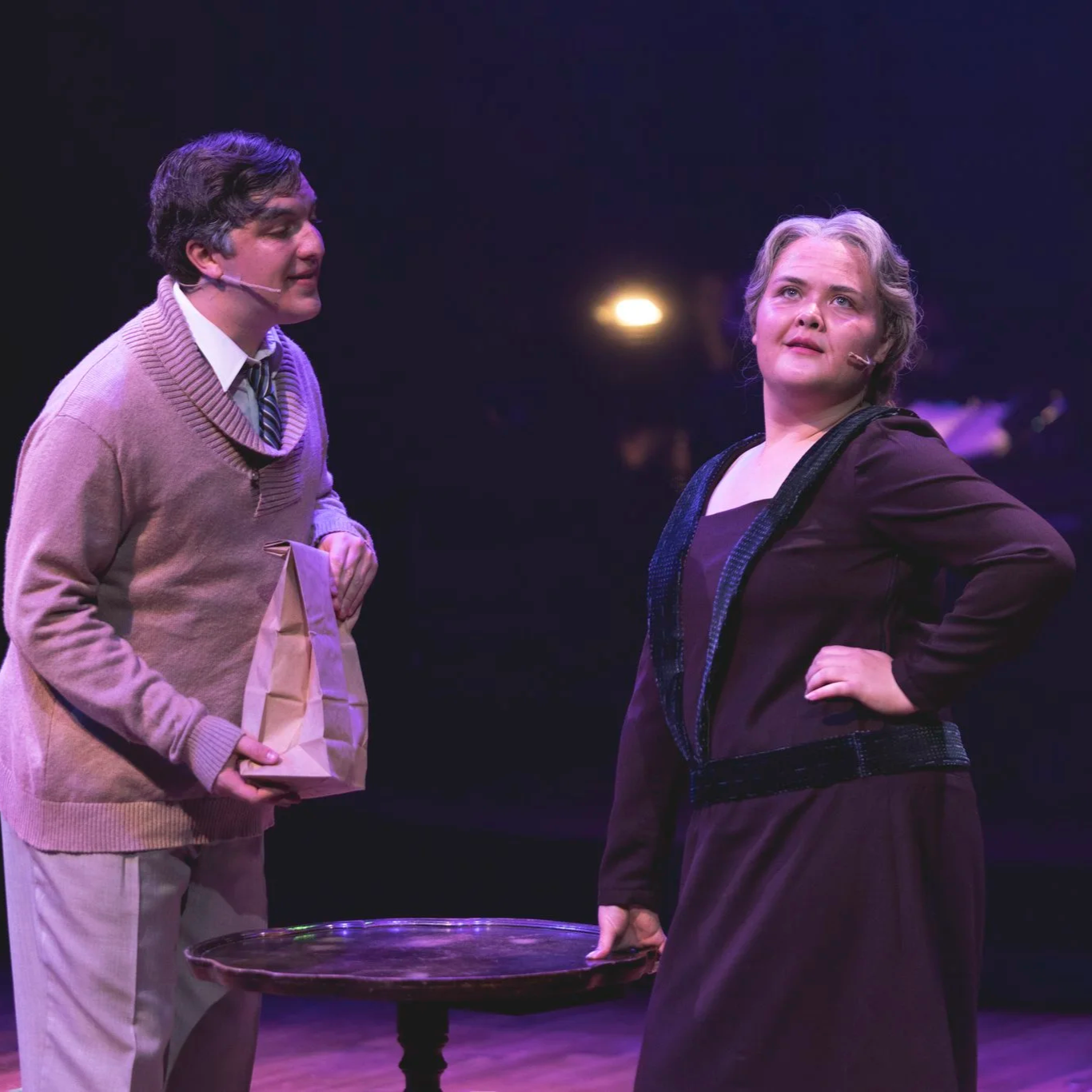 Heather is onstage in a grey wig and purple 1920s-style dress. A college-age man with light skin, greyed hair, and a beige sweater sings to her, and she gazes off with her hand on her hip.