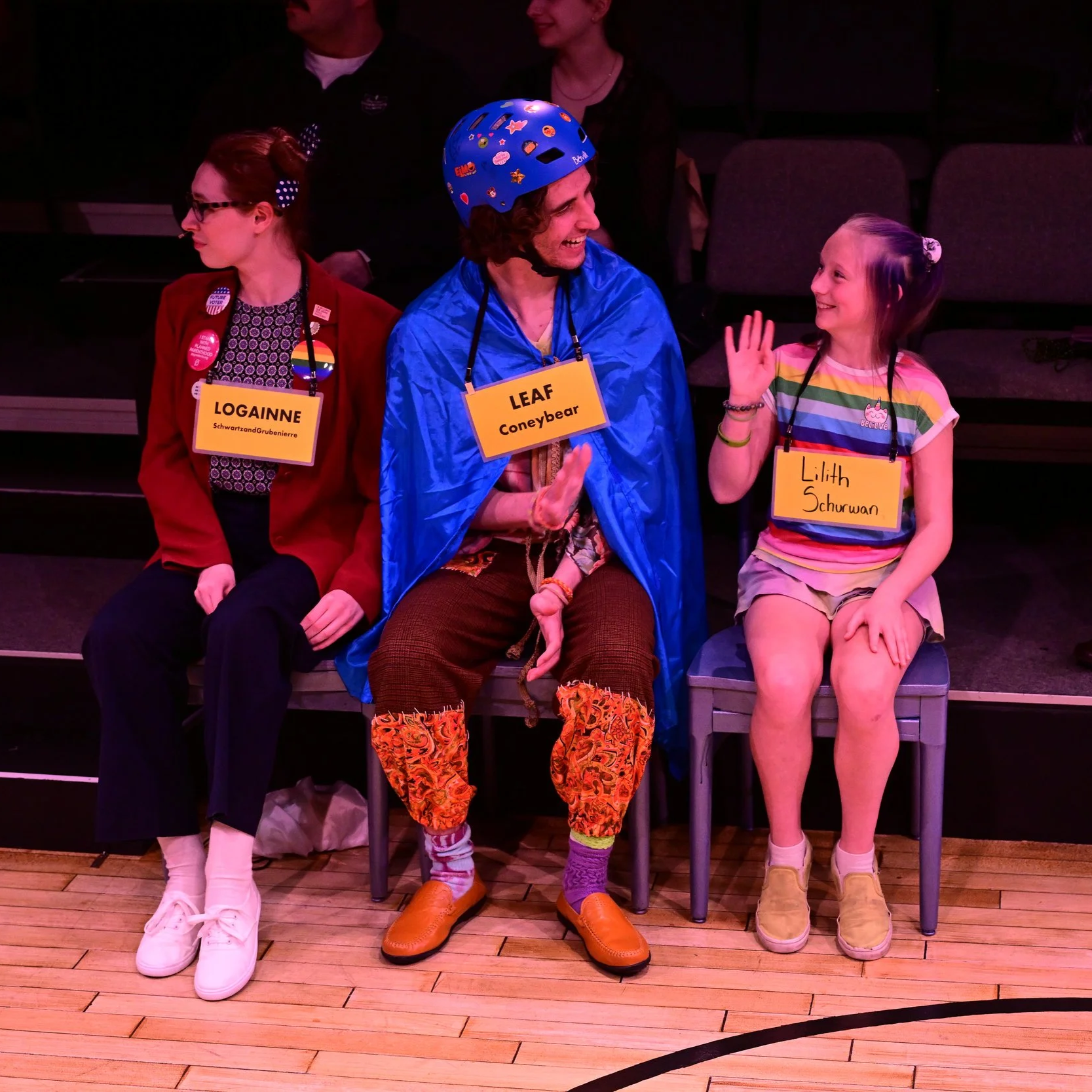 A college-age white man sits next to a child and waves excitedly. He wears a blue cape, a blue helmet covered with stickers, patchwork brown-and-orange pants with messy stitchwork, green and purple socks, and leather slip-on shoes.
