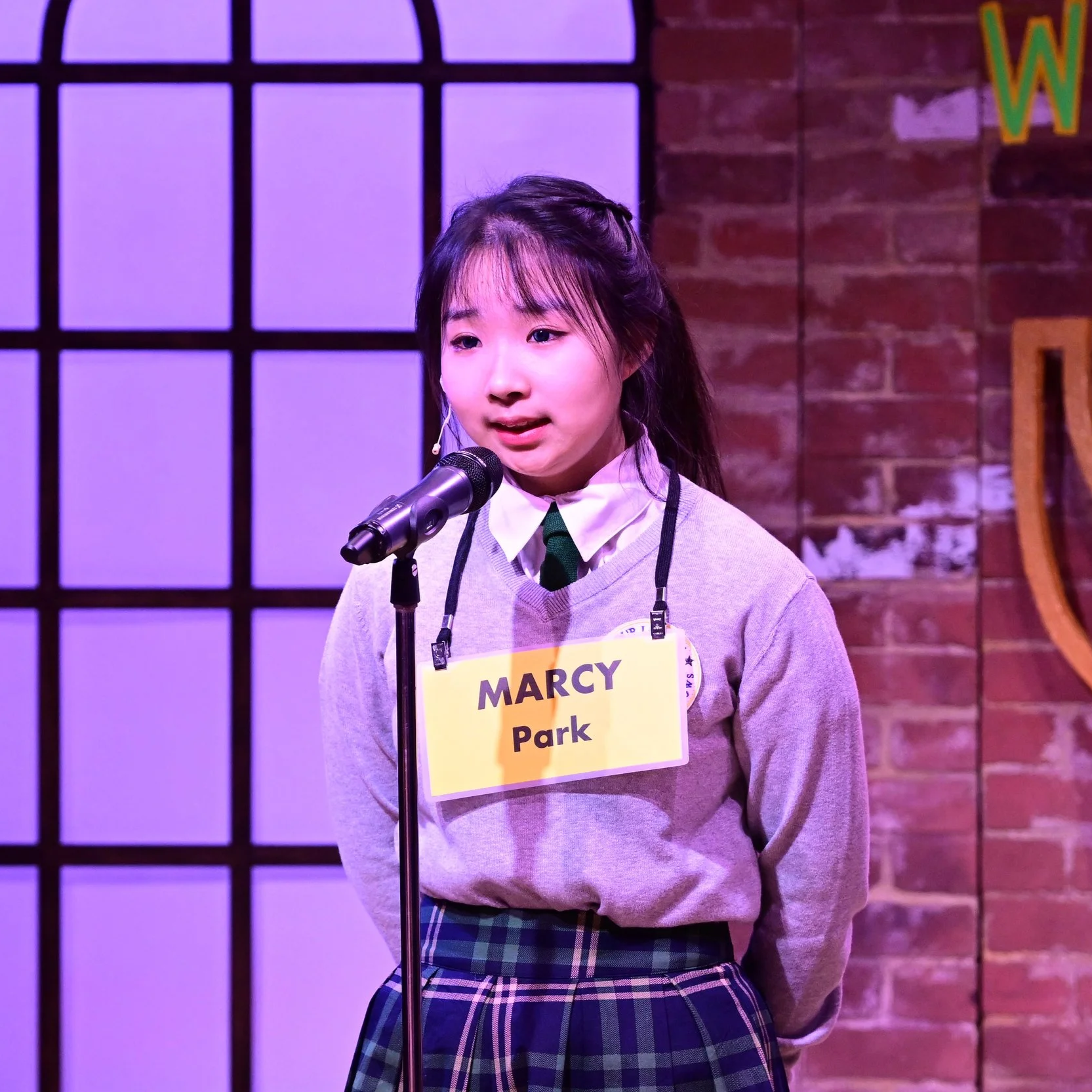 A college-age Korean woman stands in front of a microphone and speaks with her hands behind her back. She wears a grey, green, and blue boarding school uniform, and her hair is braided back.