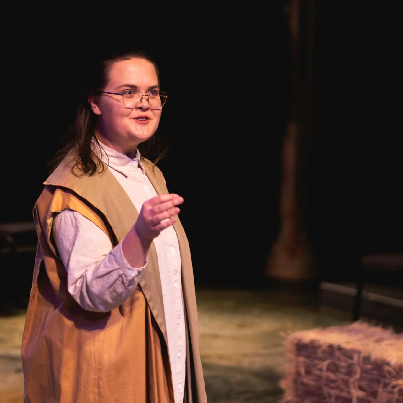 Heather is onstage wearing glasses, a rustic tan coat, and a long white tunic. With wide eyes, she speaks to an off-camera figure, gesturing confidently.