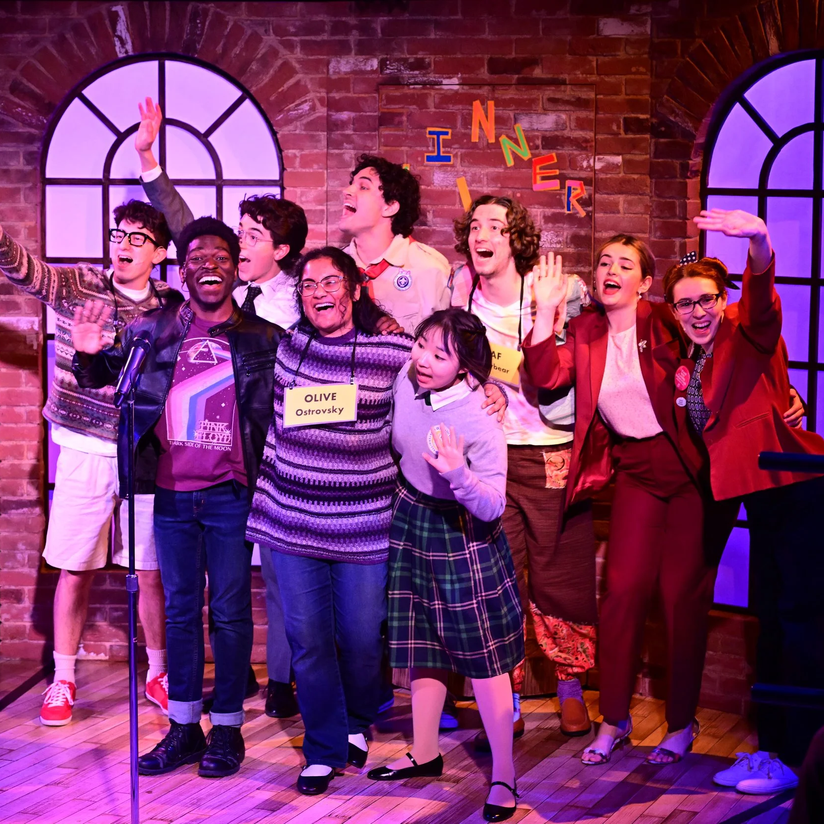The cast waves excitedly at the audience. They stand against a brick backdrop with tall, rounded windows.