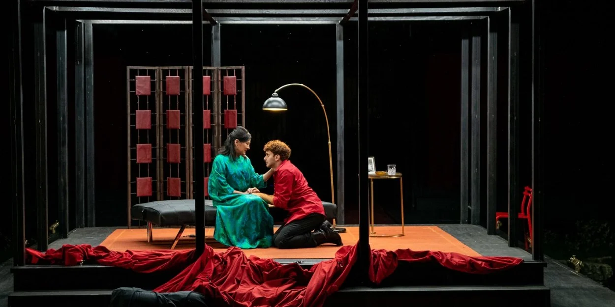 A light-skinned Middle Eastern man in his 30s wears a red buttoned shirt and kneels in front of a middle-aged Japanese woman in a teal 1950s dressing gown. She puts her hand on his cheek.