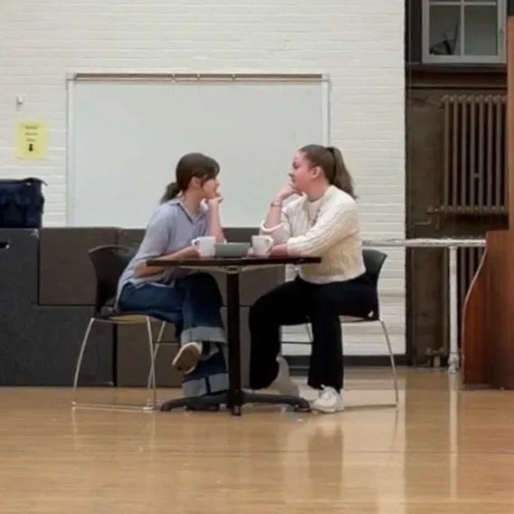 Two college-age white women sit across from each other at a small table. They rest their hands on their chins, smiling passive-aggressively at each other.