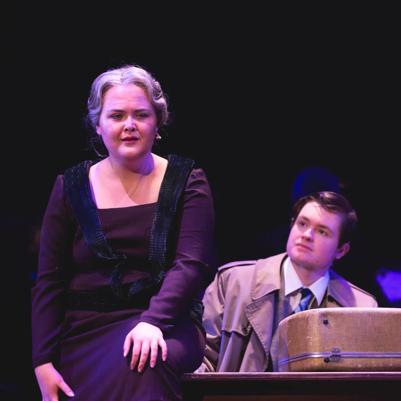 Heather is onstage in a grey wig and purple 1920s-style dress. She sings meditatively, as a white college-age man in a beige trenchcoat sits behind, listening.