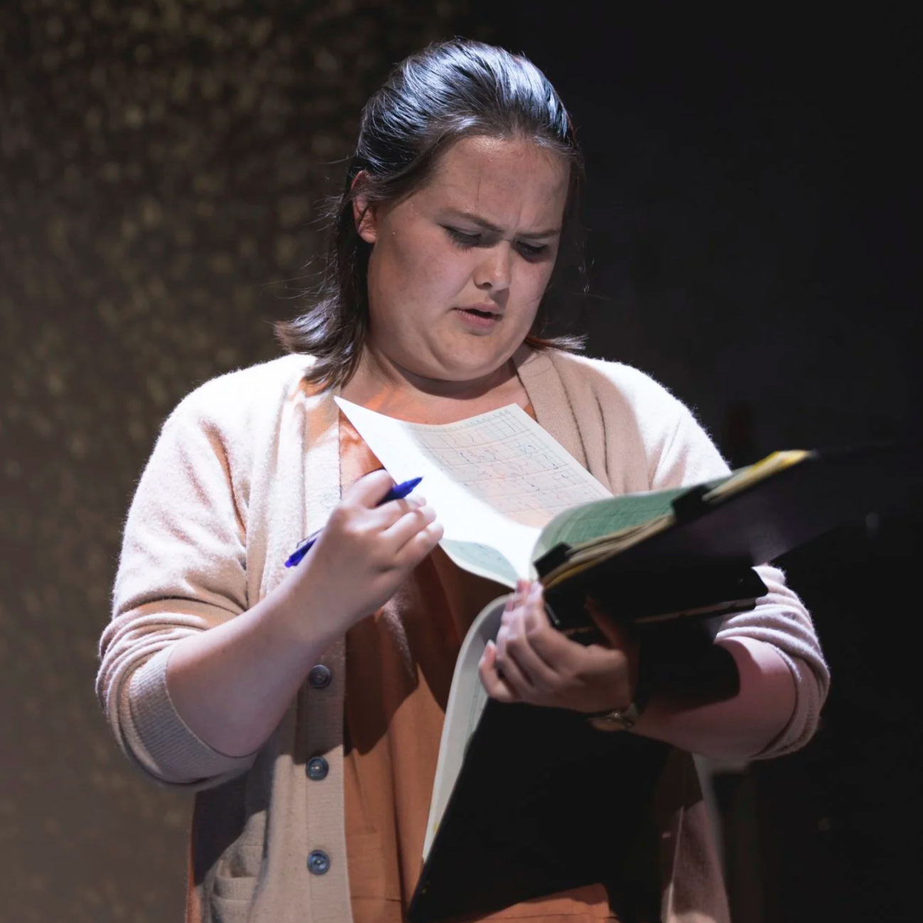 Heather is onstage in an orange dress and beige sweater. Pen in hand, she looks intently through the figures on a legal notepad and narrows her eyes.
