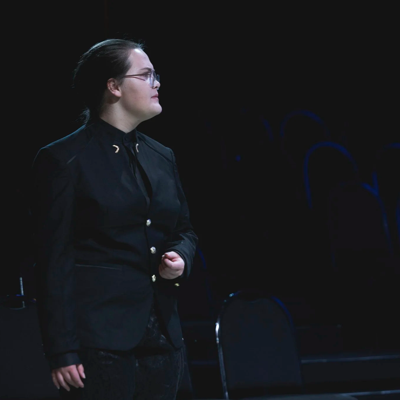 Heather is onstage, wearing glasses, a French twist, and a black suit with gold buttons and insignia. She stands tall, staring down an off-camera figure with poise. 