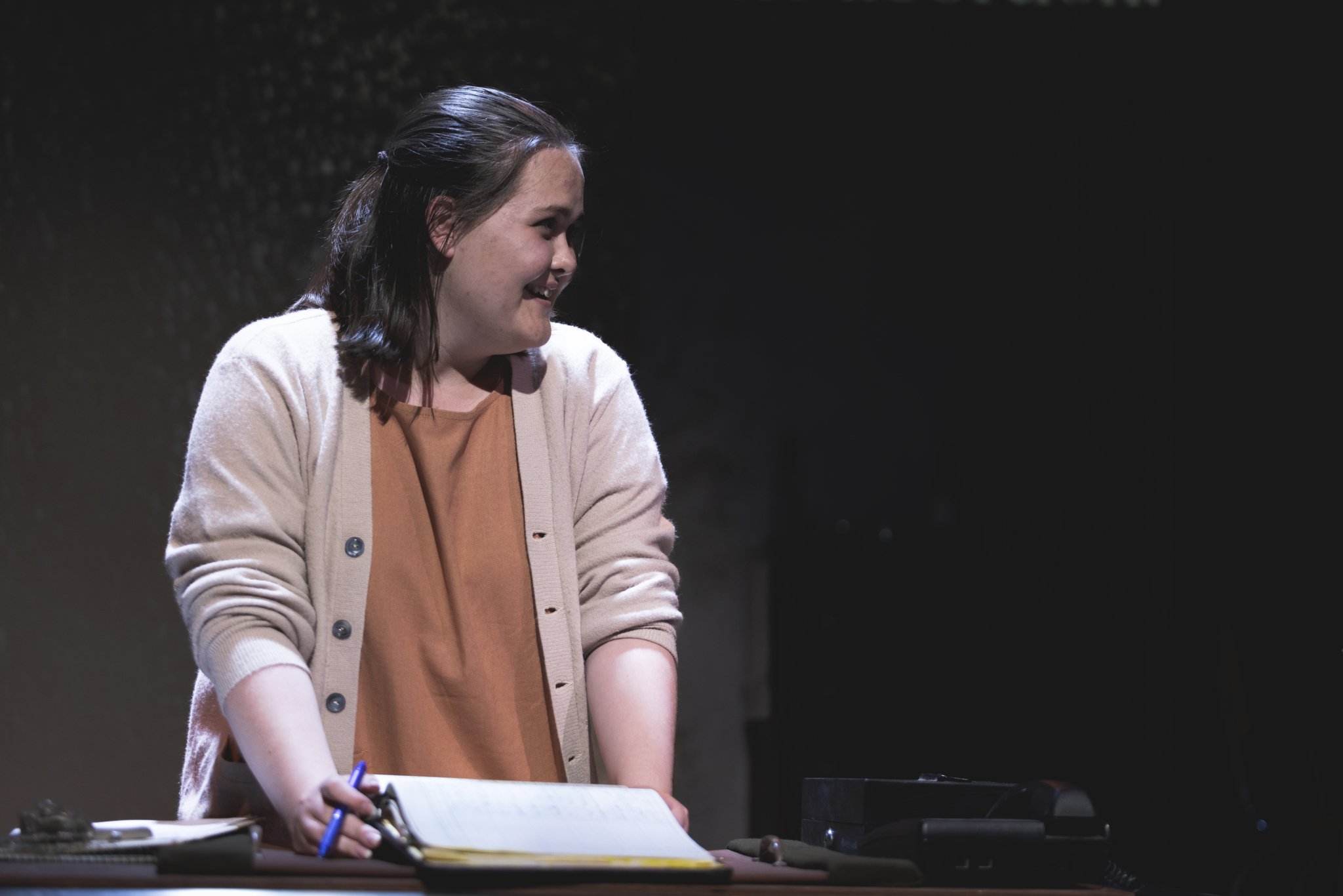 Heather is onstage in an orange dress and beige sweater. She stands at a desk and holds a pen and legal notepad. She speaks smilingly to an off-camera figure.