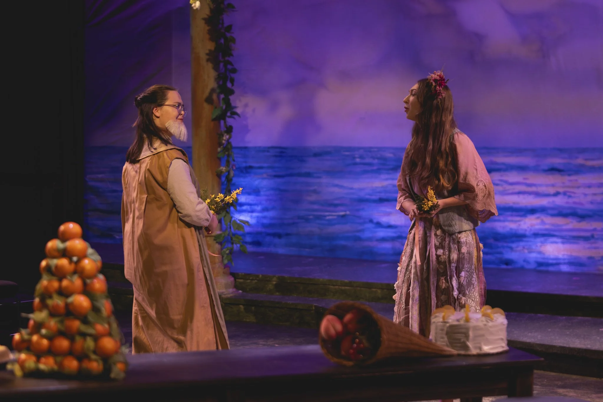 Heather is onstage holding yellow flowers. She wears glasses, a rustic tan coat, a long white tunic, and a fake white beard. She looks at a college-age white woman in a flowing pink dress, bemused and intrigued.