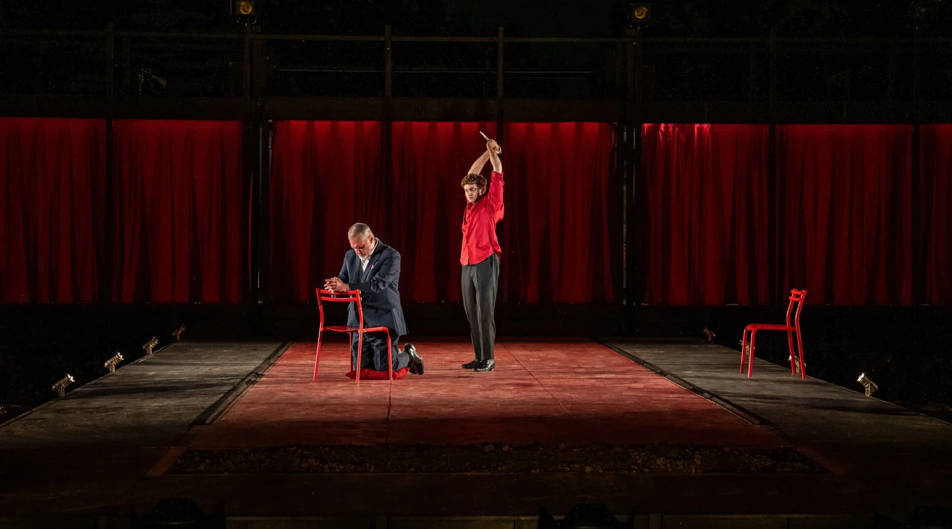 A light-skinned Middle Eastern man in his 30s wears a red buttoned shirt and raises a dagger above his head. He looks furiously at his target, a middle-aged white man in a suit who kneels to pray.