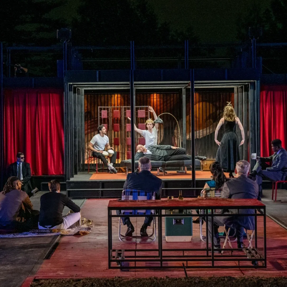 The company sits on a stark, red-and-black stage framed by red curtains. They watch a group of actors perform an experimental, Martha Graham-style dance.