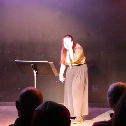 Heather is onstage beside a music stand. Her hair is half-up half-down, and she wears a green buttoned shirt with flowing black pants. She covers one eye with her hand, staring in shock.