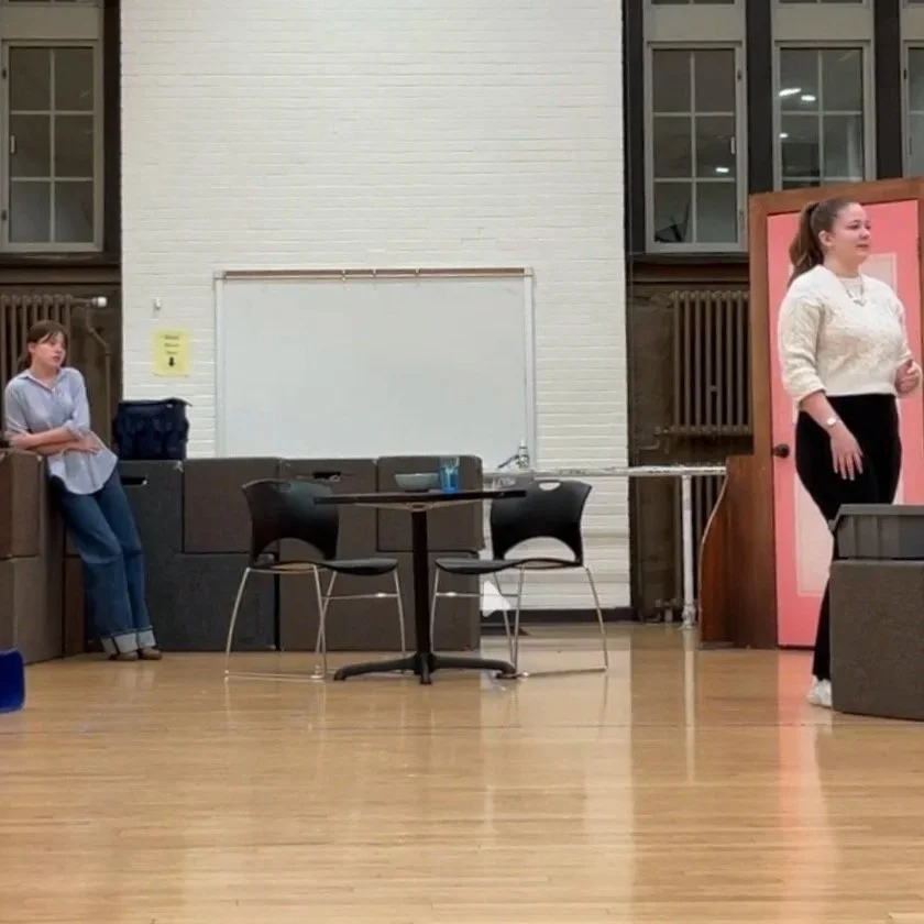 A college-age white woman in a white blouse speaks meditatively. Behind, a college-age white woman in a striped blue shirt leans against acting blocks, watching her.