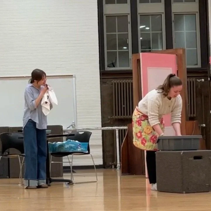 A college-age white woman holding a bloody rag looks at another college-age white woman in a pink flowered apron, who roots through a drawer.