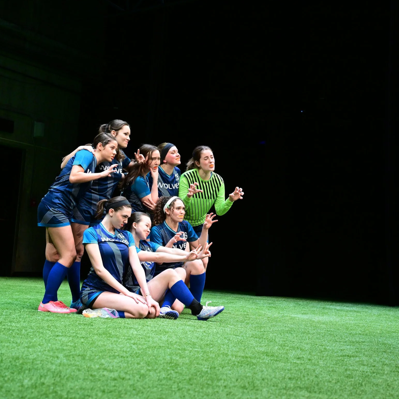 Young AFAB soccer players in dark blue uniforms stand on a pitch with shadowy lighting. They make silly poses for the camera, with orange slices in their mouths.