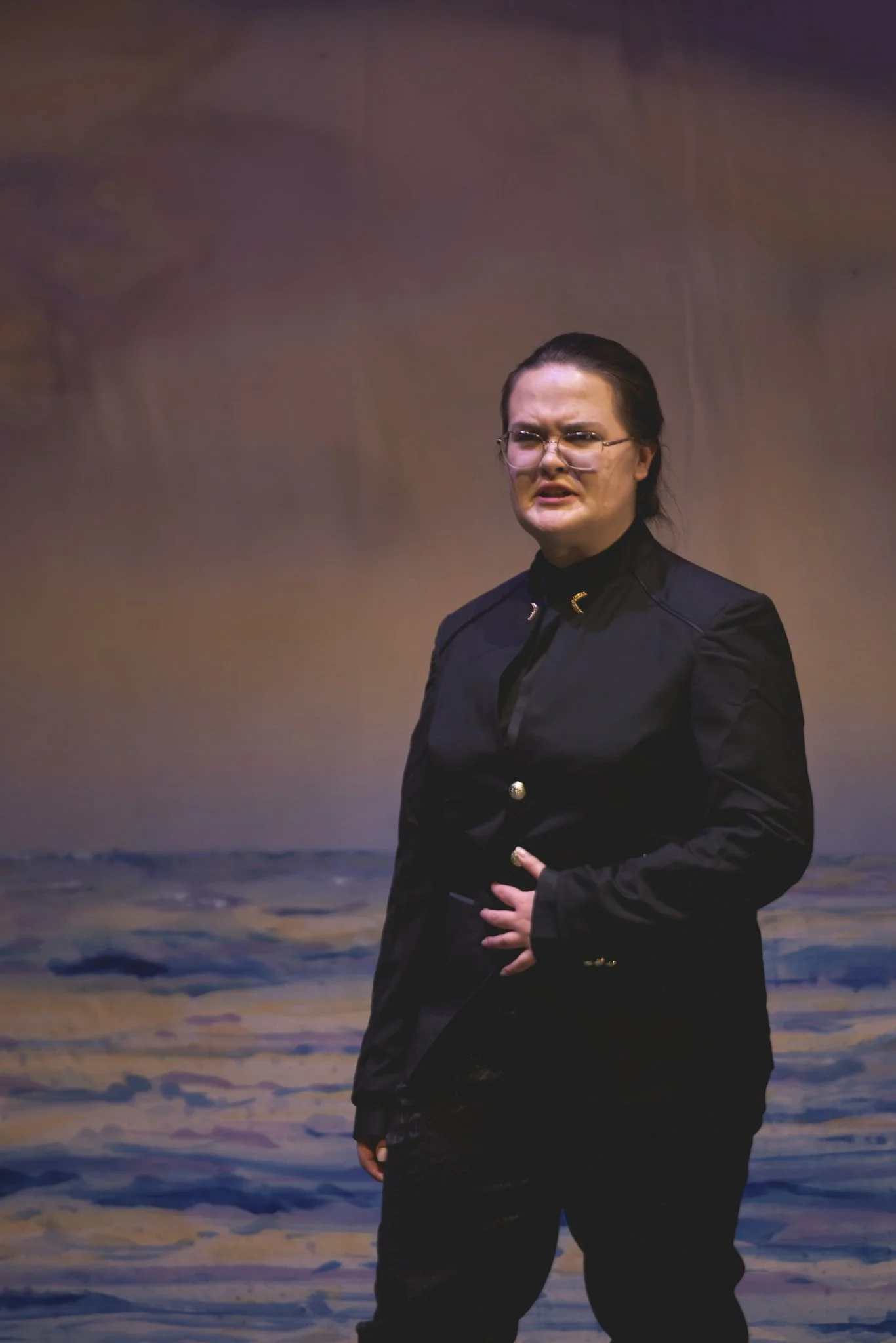 Heather is onstage, wearing glasses, a French twist, and a black suit with gold buttons and insignia. She puts her hand to her stomach and speaks with disgust.