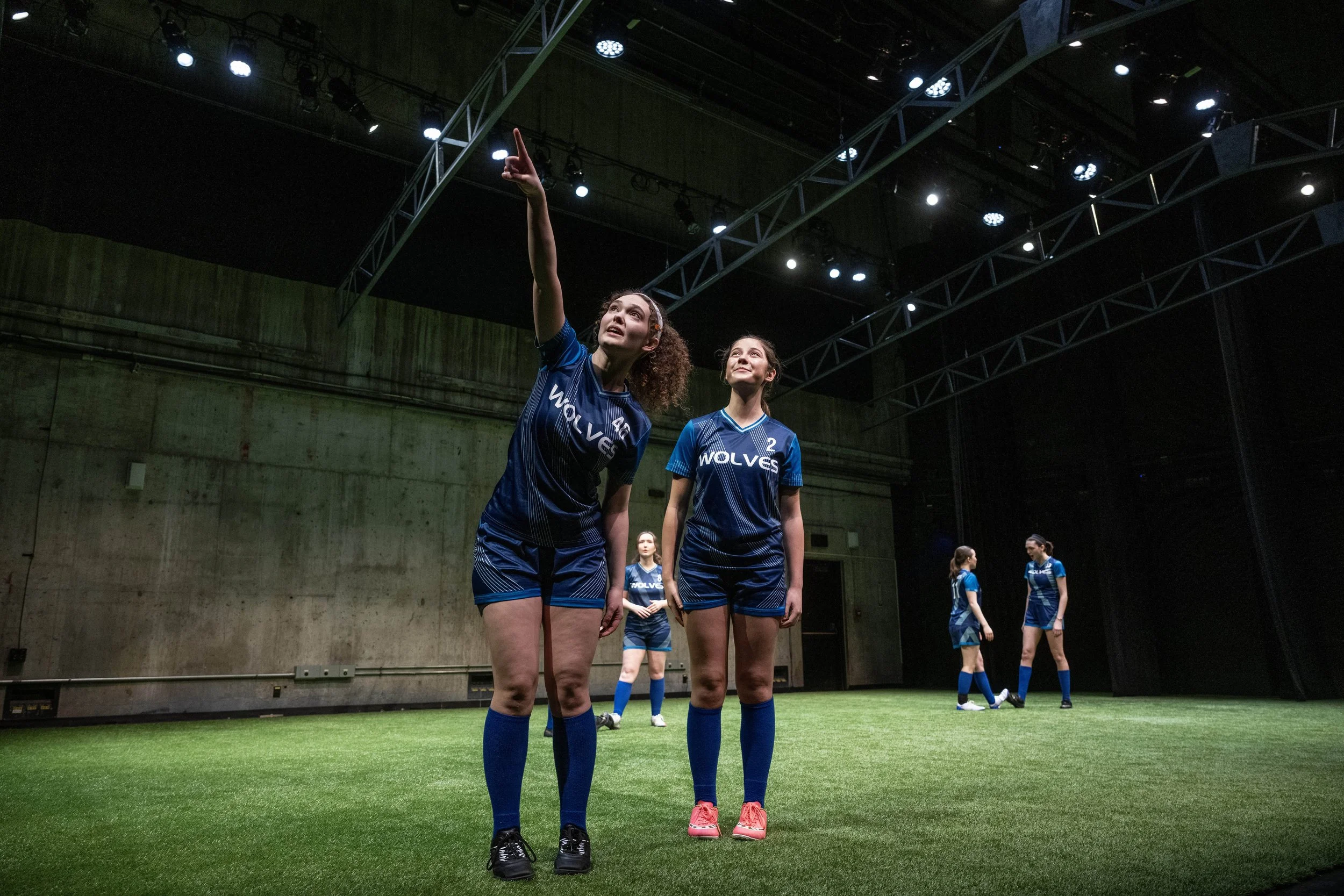 Young AFAB soccer players in dark blue uniforms stand on a pitch with shadowy lighting. One player points to the ceiling, and another player looks where she points.