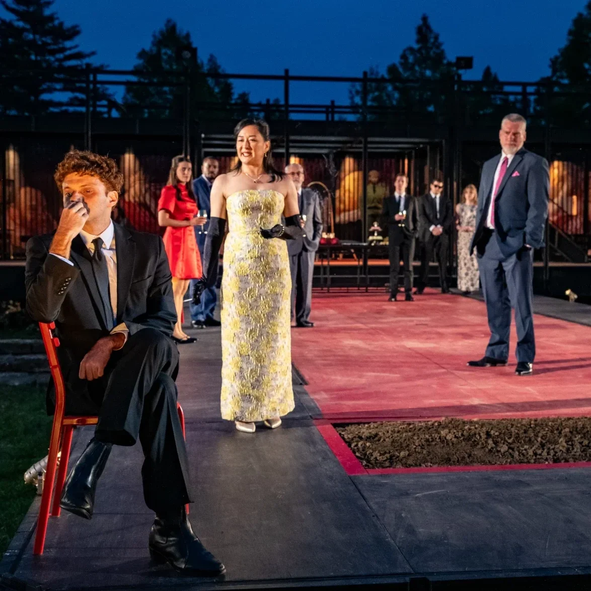 A light skinned, Middle Eastern man in his 30s wears a black suit and sits in a chair. He looks sadly into the distance as a middle-aged Japanese woman in a gold 1950s gown speaks to him. The company stands behind, looking at him.
