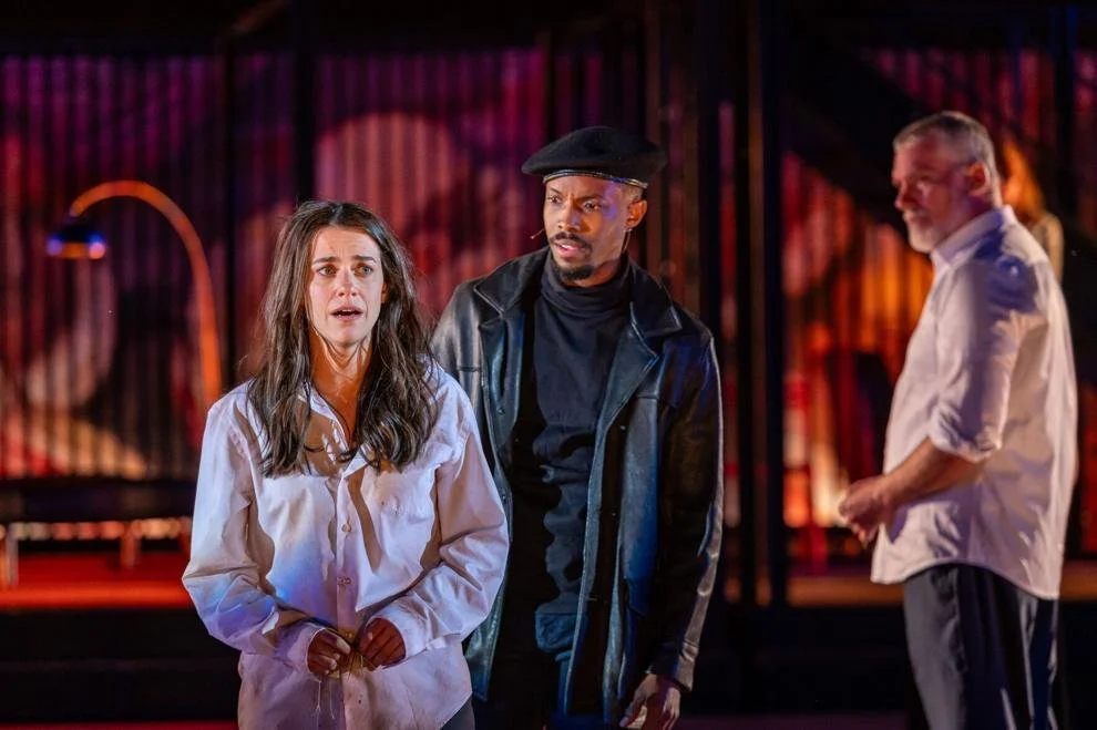 A white woman in her 30s speaks distressedly. She wears a wrinkled white buttoned shirt, and her long, brown hair is tussled. A Black man in his 30s stands behind her, watching with horror. He wears a black leather jacket and beret.