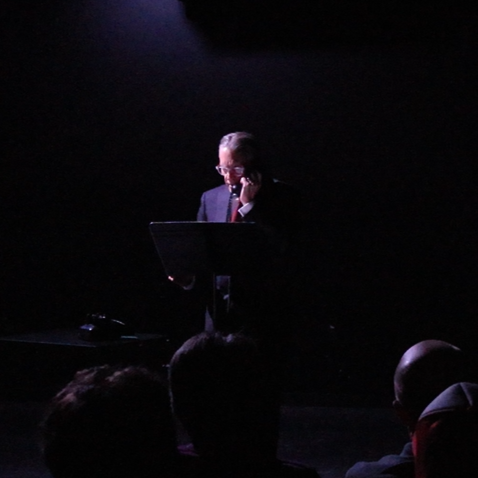 A white middle-aged man is in front of a music stand, holding a black dial telephone receiver to his ear. His white hair is slicked back, and he wears glasses and a blue suit.