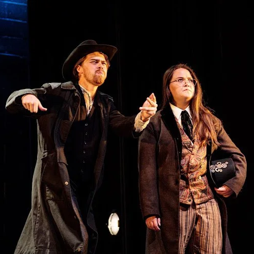 Heather is onstage in a brown wool peacoat and a patterned vest. She holds a black top hat with gears, and stares off frustratedly as a white actor in a black trenchcoat and cowboy hat jumps and flails.