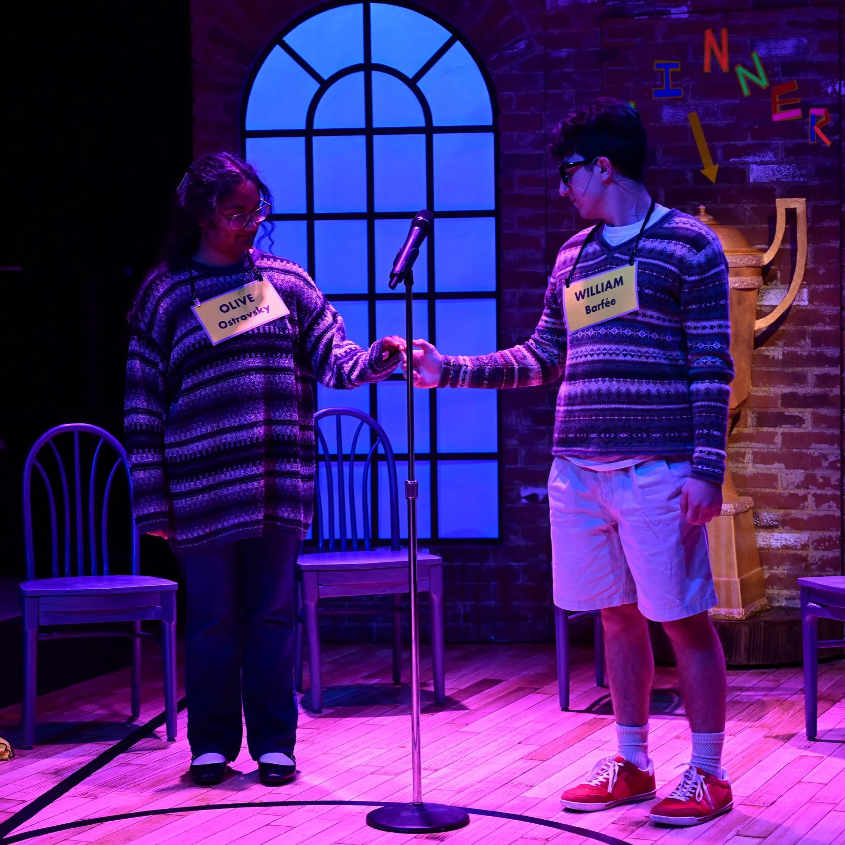 A college-age Indian woman stands onstage in a striped purple and black sweater. She holds her hand out to a college-age white man in a yellow striped argyle sweater and thick glasses. The lighting is purple and tender.