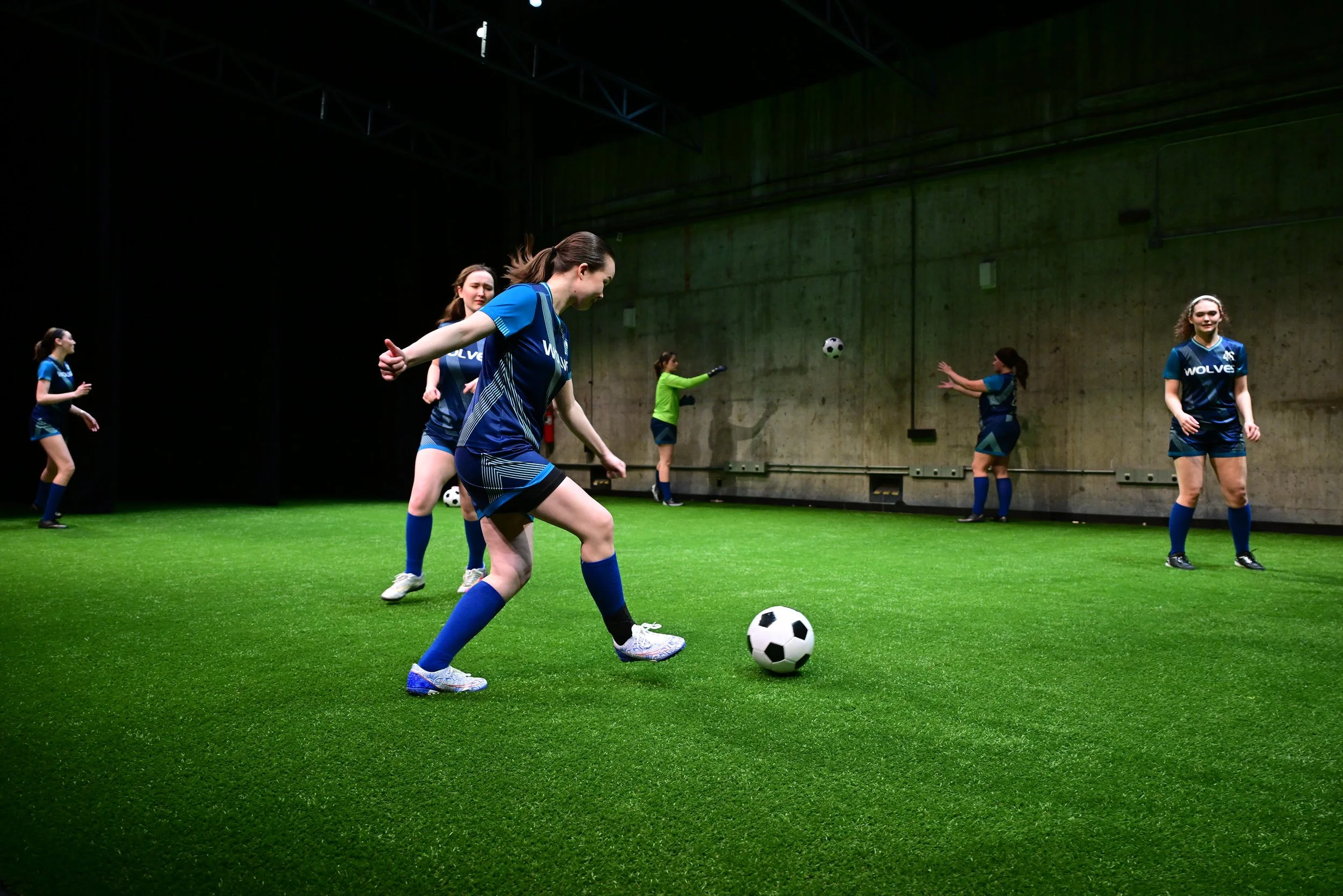 Young AFAB soccer players in dark blue uniforms stand on a pitch with shadowy lighting, running drills. They kick and throw soccer balls.
