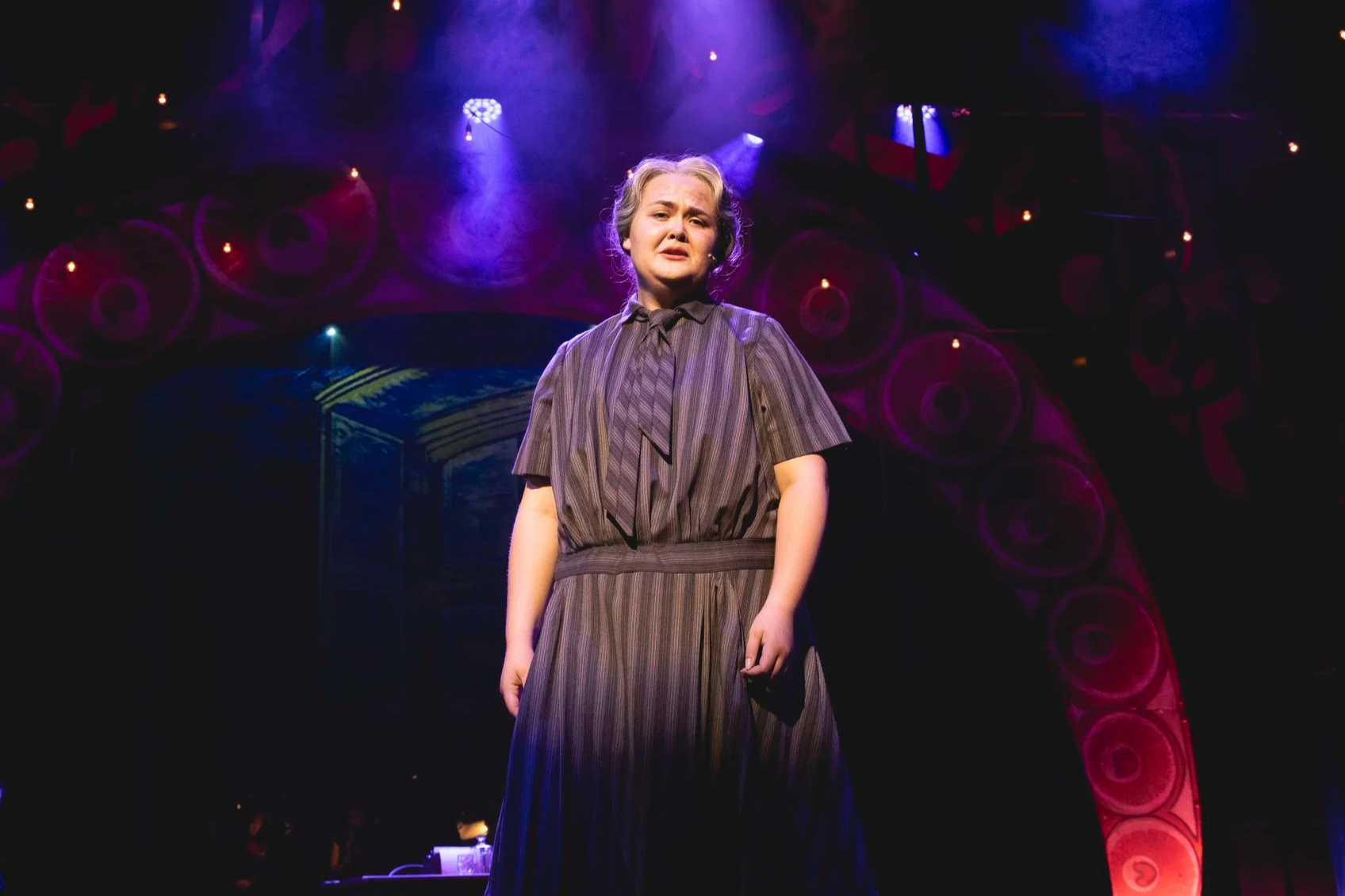 Heather is onstage in a grey wig and a grey striped dress. She looks directly at the viewer and sings sadly. Lights dimly illuminate the stage fog behind her.